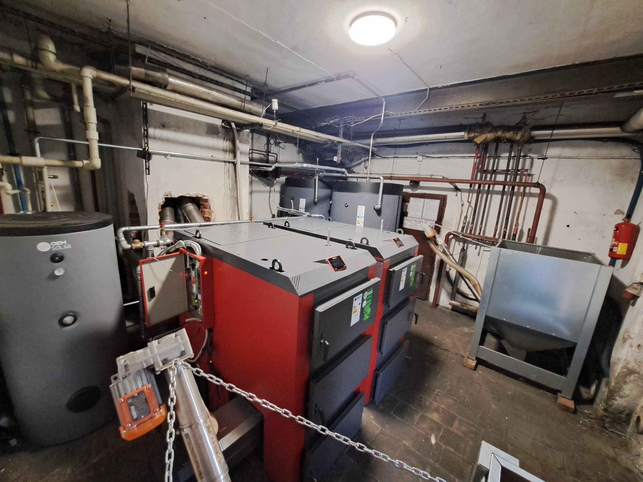 Dwa kotły grzewcze z podajnikiem w kotłowni, widoczne rury instalacji, zbiornik buforowy i podajnik. Oświetlenie sufitowe i ceglana podłoga.