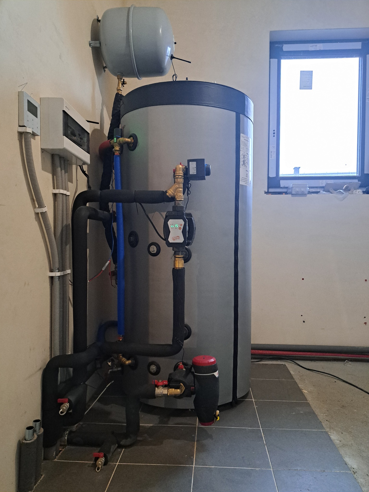 Szary zbiornik buforowy z osprzętem i rurami w kotłowni, widoczne elementy instalacji grzewczej, okno w tle. Ujęcie z niskiego kąta, podkreślające wysokość zbiornika.