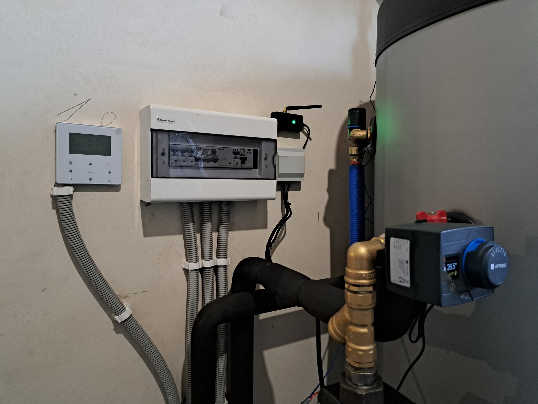 Szara instalacja grzewcza z czarnymi rurami, białą skrzynką elektryczną i regulatorem temperatury Afriso na tle jasnej ściany. Widoczne przewody i elementy montażowe.