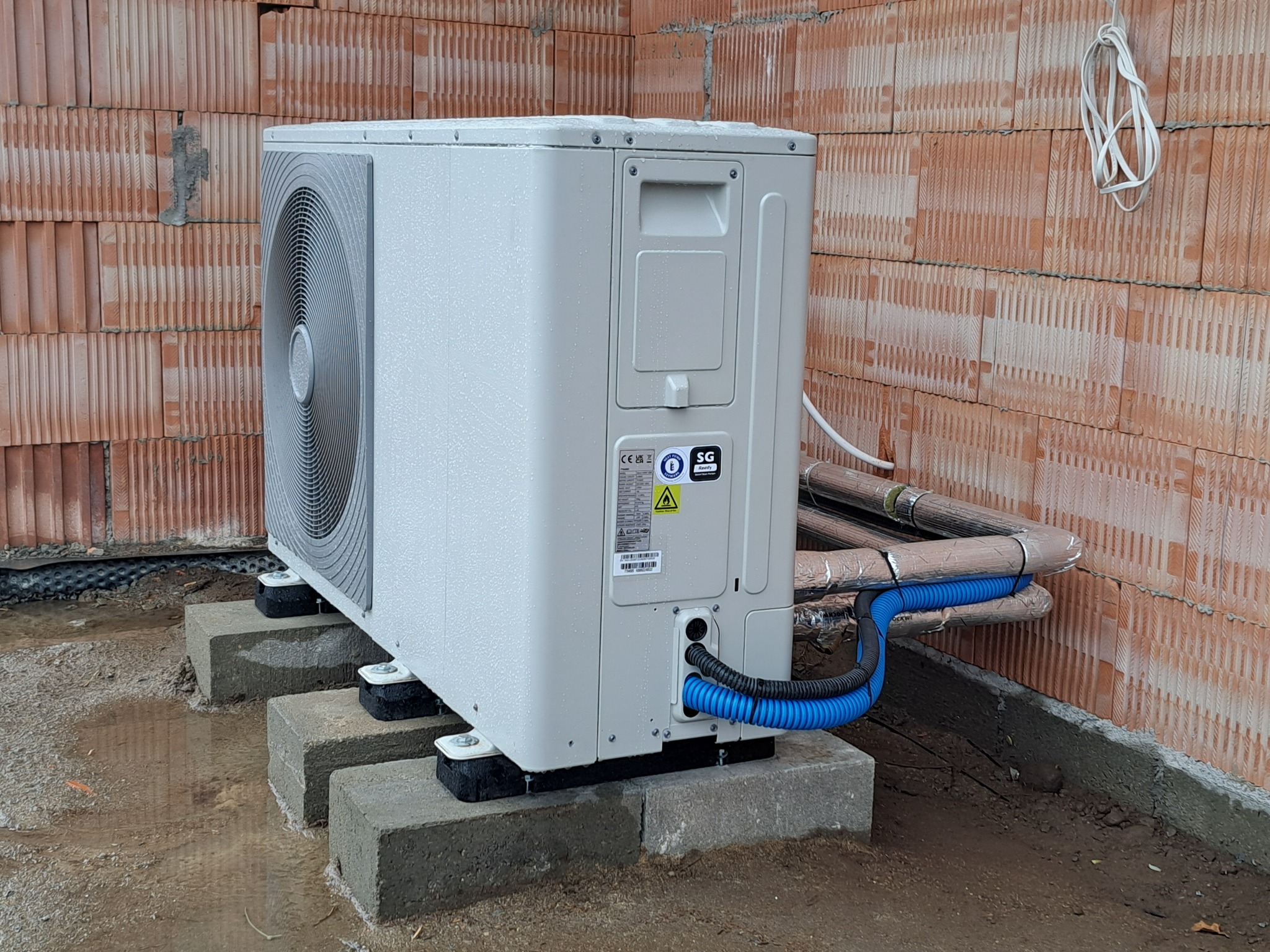 Biała pompa ciepła na betonowych blokach, z widocznymi rurami i przewodami, zainstalowana przy ścianie z czerwonej cegły. Widoczne ślady wilgoci na podłożu.