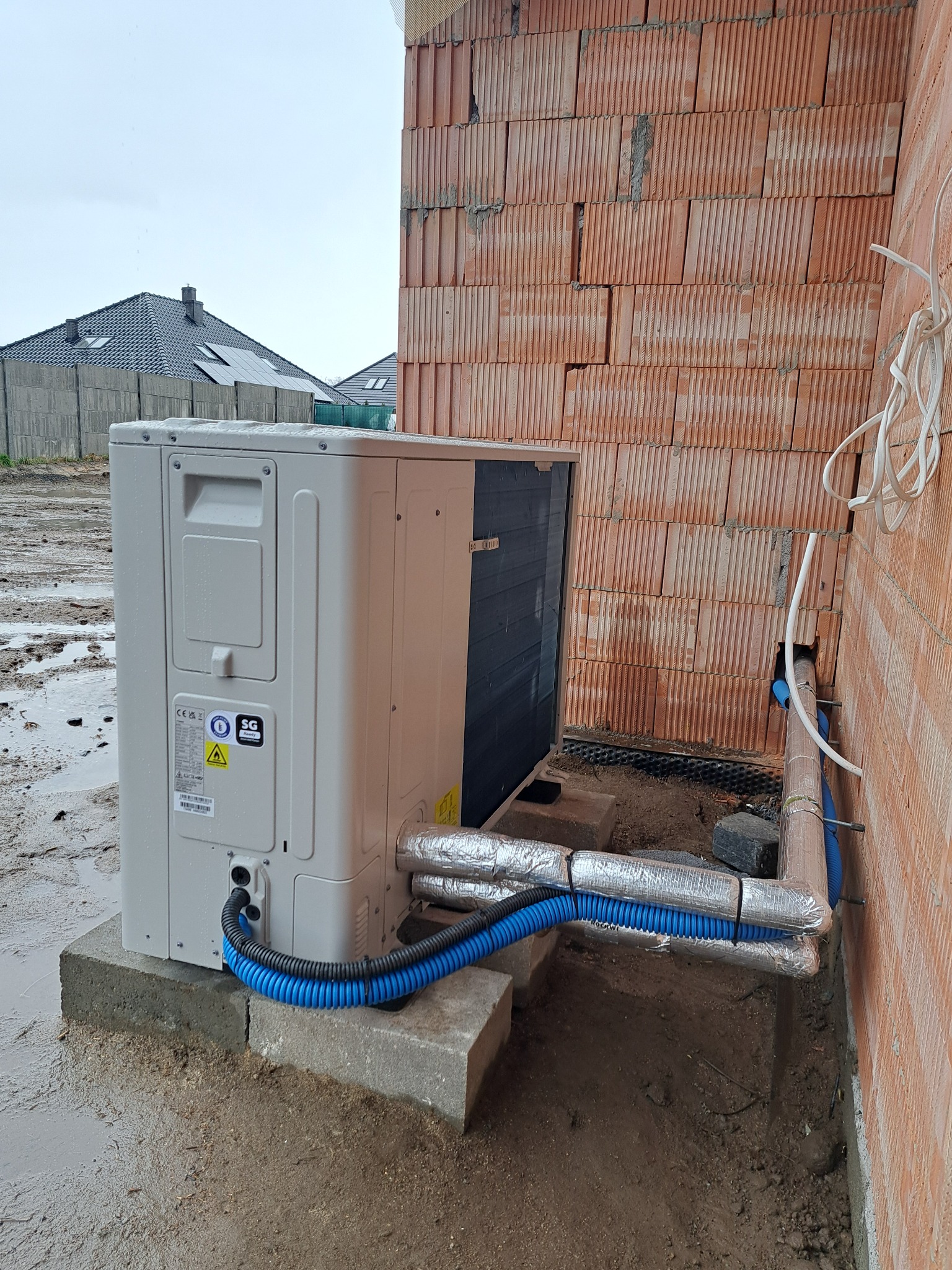 Biała pompa ciepła zamontowana na betonowym fundamencie przy ścianie z czerwonej cegły. Widoczne niebieskie rury i srebrna izolacja termiczna. W tle domy i pochmurne niebo.