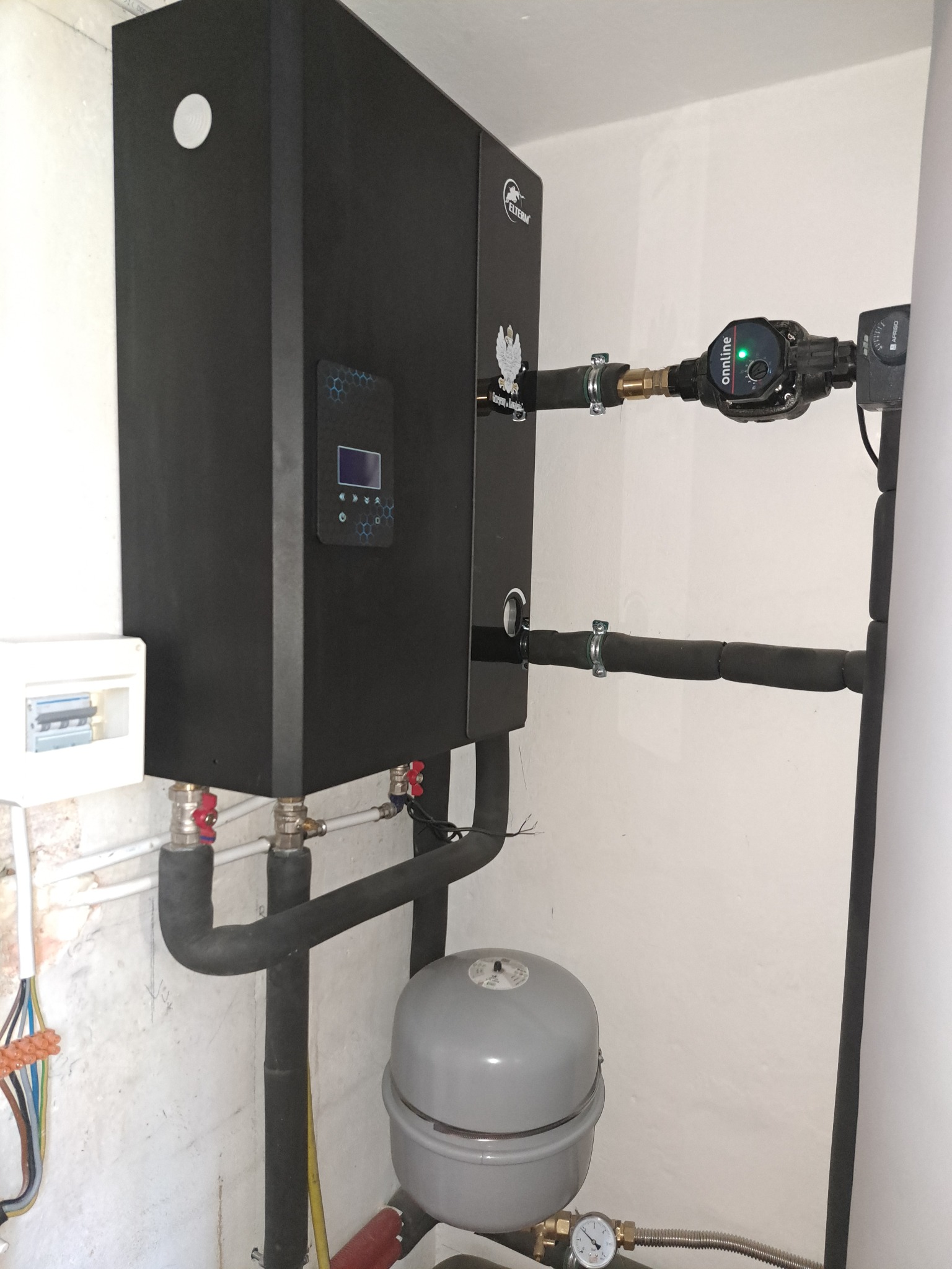 Czarny piec z wyświetlaczem i orłem na obudowie, podłączony do instalacji z rurami w izolacji, zamontowany na białej ścianie w pomieszczeniu.