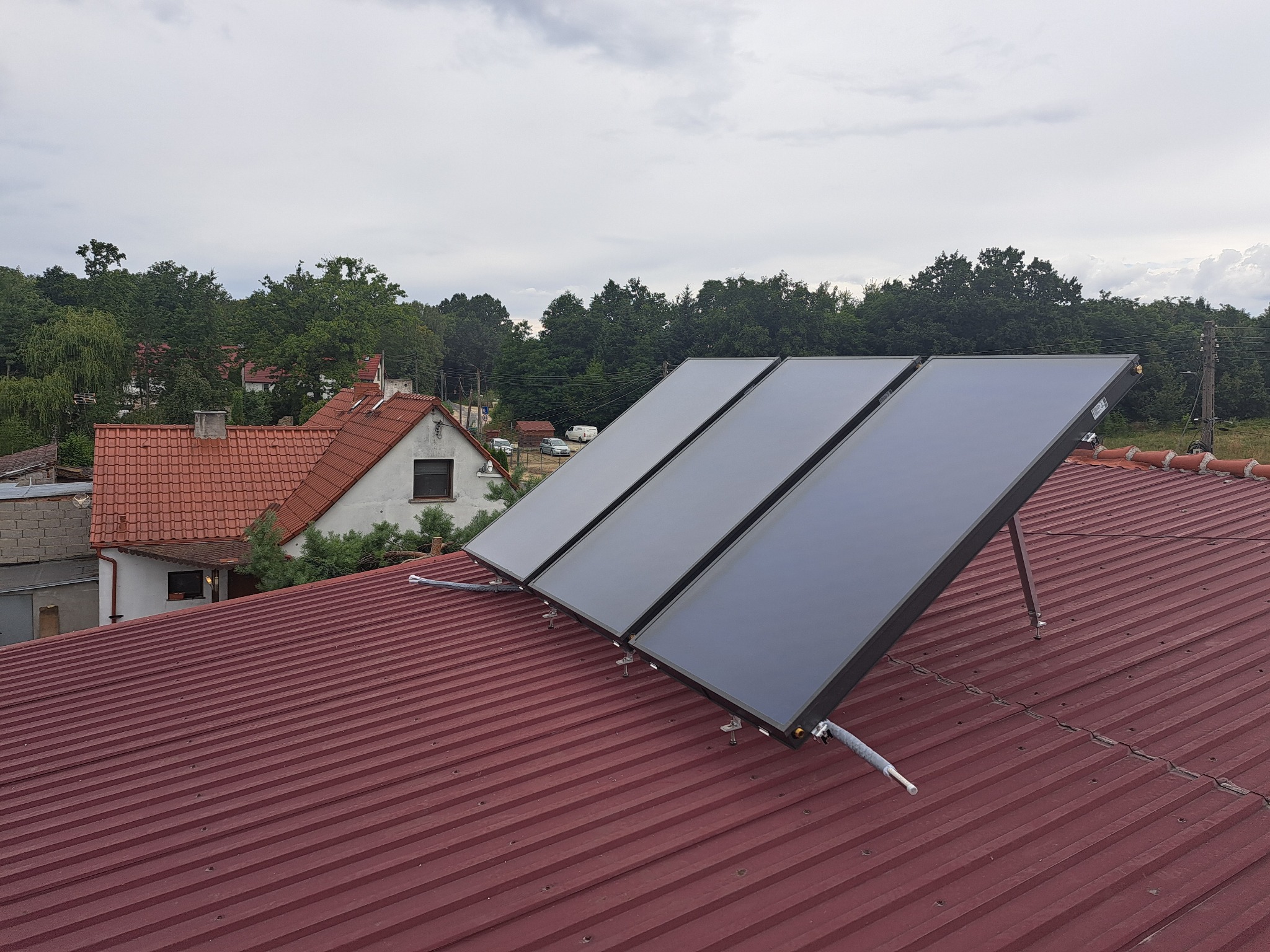 Trzy panele słoneczne zamontowane na czerwonym, blaszanym dachu domu jednorodzinnego. W tle widoczne drzewa i zabudowania.
