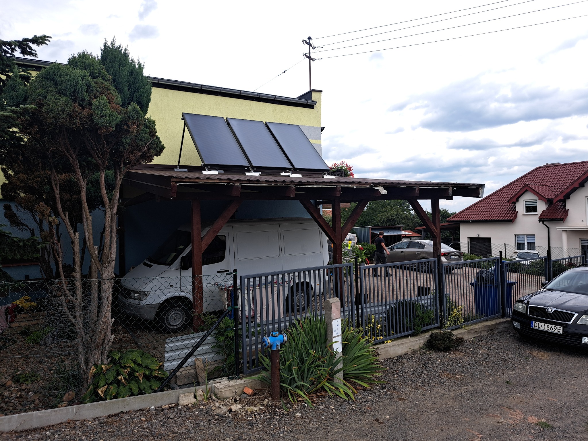 Instalacja solarna na drewnianej wiacie garażowej przy żółtym budynku. Biały van zaparkowany wewnątrz, widoczne ogrodzenie i samochody w tle.