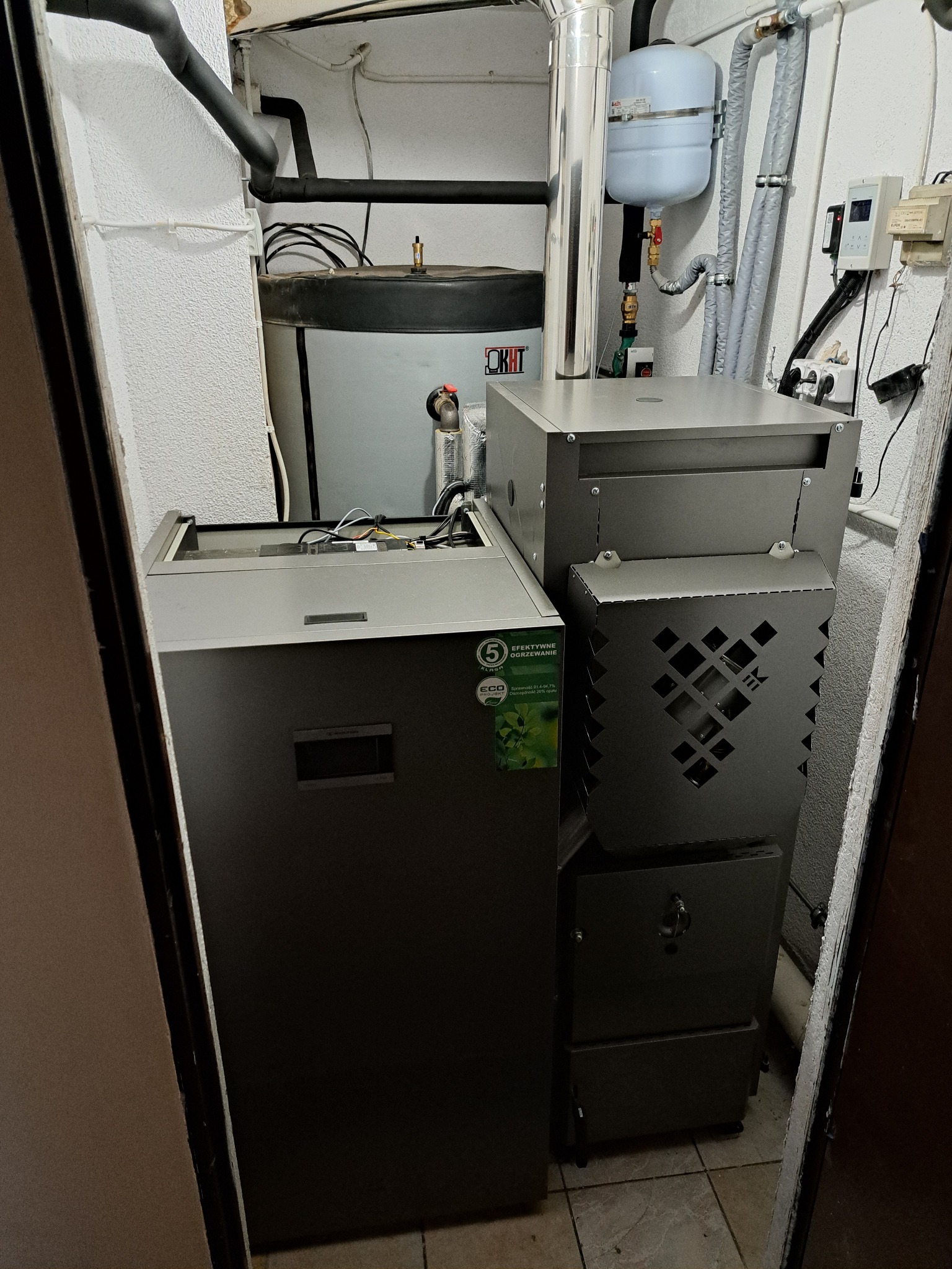 Szary piec CO z podajnikiem w kotłowni, widoczne rury i zbiornik wyrównawczy. Piec z certyfikatem klasy 5 i oznaczeniem ECO. Ujęcie z bliska, w ciasnym pomieszczeniu.