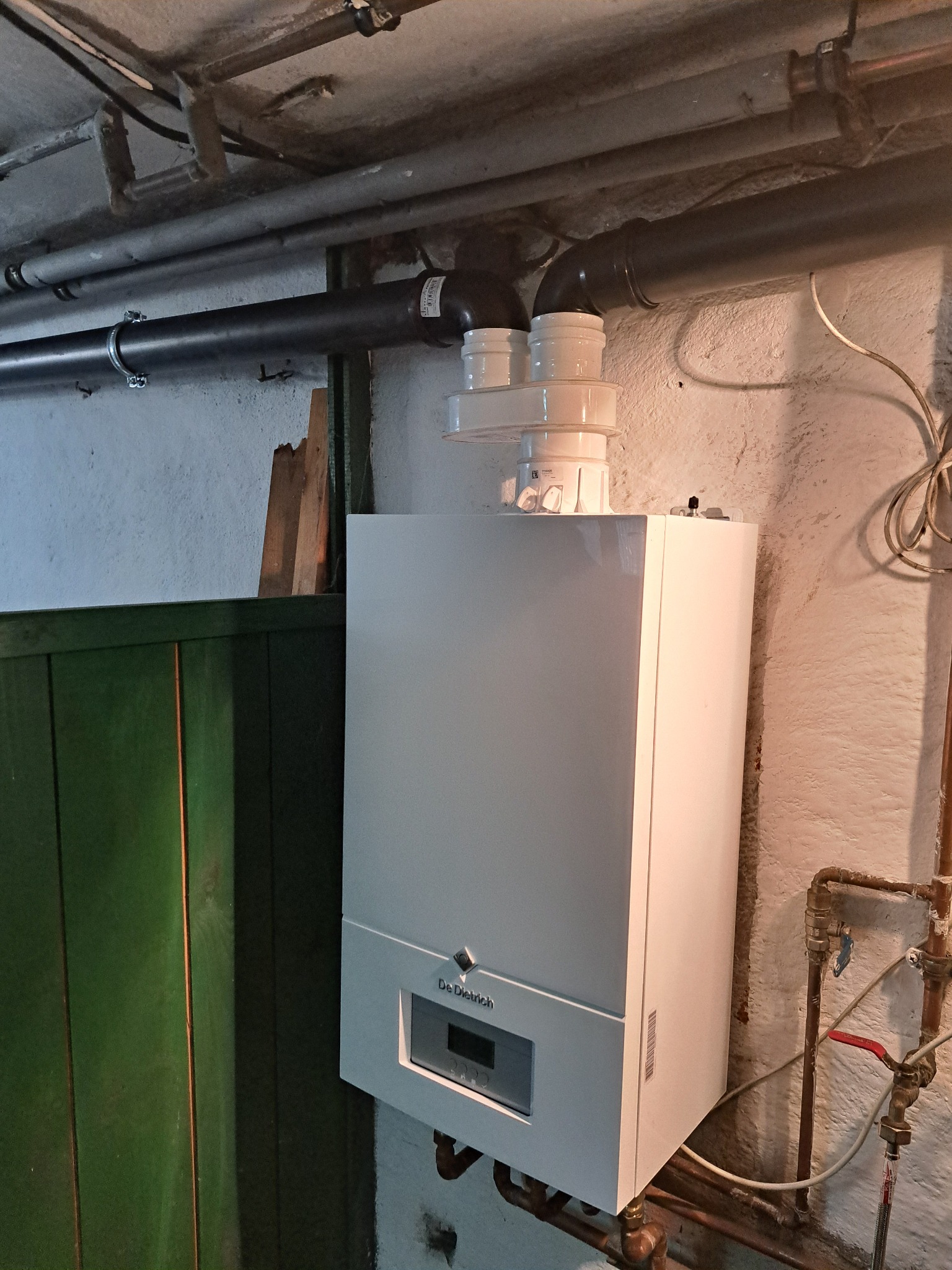 Biały piec gazowy De Dietrich z podłączeniami w piwnicy, widoczne rury i zawory. Ściana z surowym wykończeniem, obok zielone drzwi.