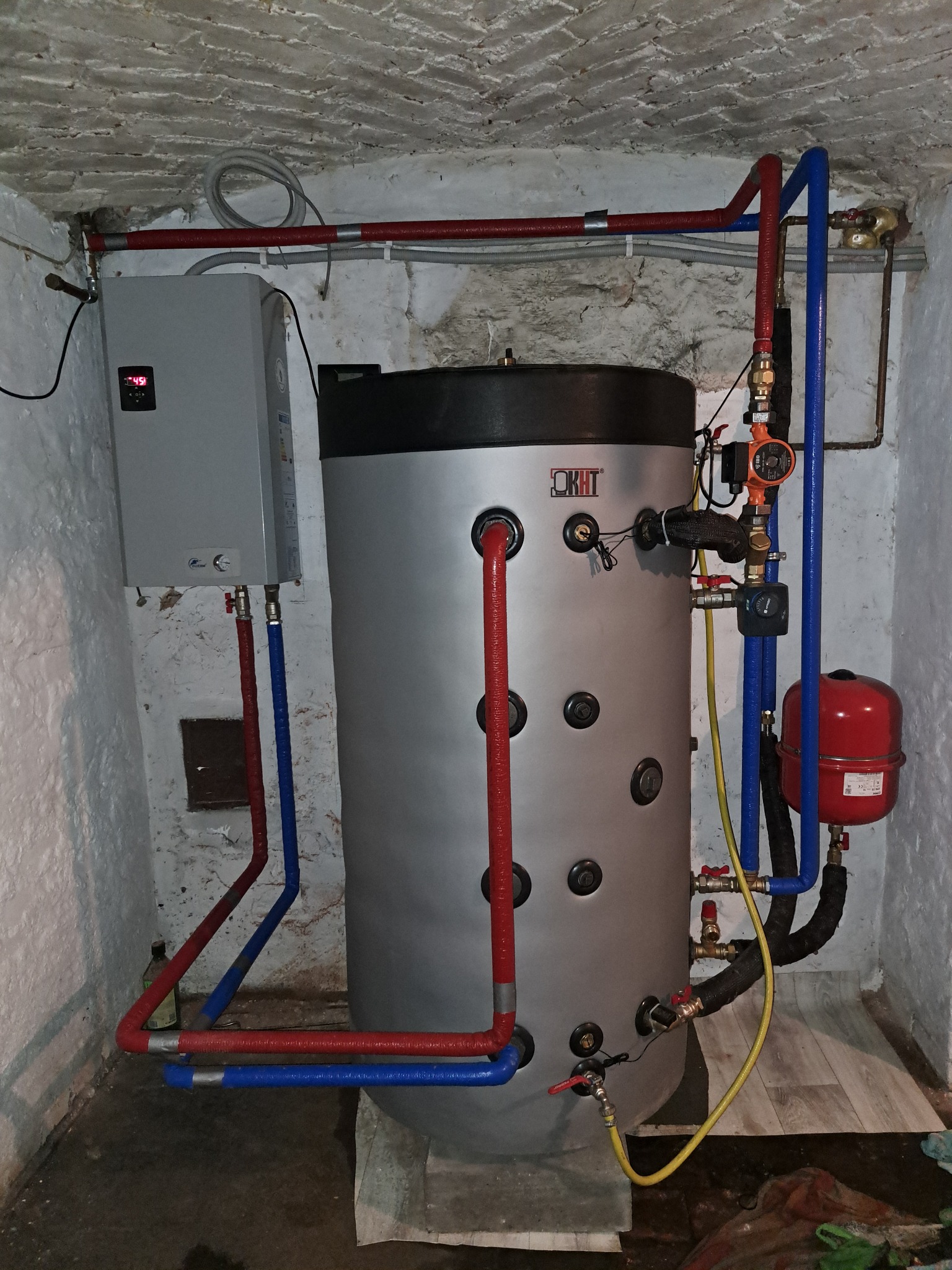Zamontowany zasobnik ciepłej wody użytkowej OKHT z podłączonymi rurami w kolorach czerwonym i niebieskim, oraz dodatkowymi elementami instalacji w piwnicy.