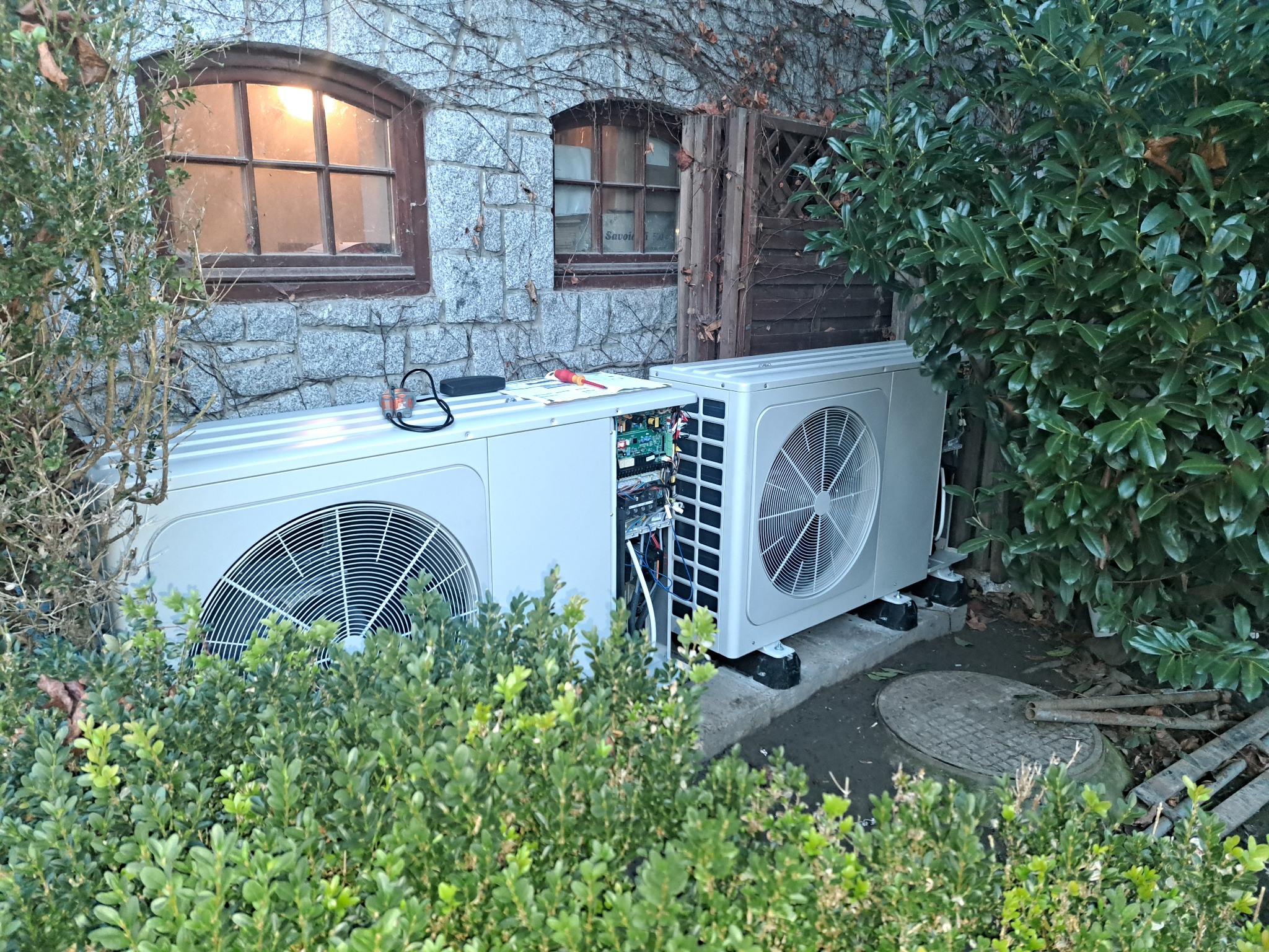 Dwie jednostki zewnętrzne pompy ciepła, jedna z otwartą obudową, widoczne okablowanie, na tle kamiennej ściany budynku z oknami i zielenią.