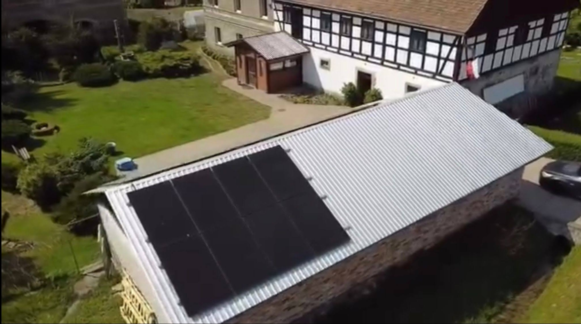 Panele słoneczne na dachu budynku w Legnicy, widok z góry. Cztery czarne panele na srebrnym, blaszanym dachu. W tle budynek z muru pruskiego i zielony trawnik.