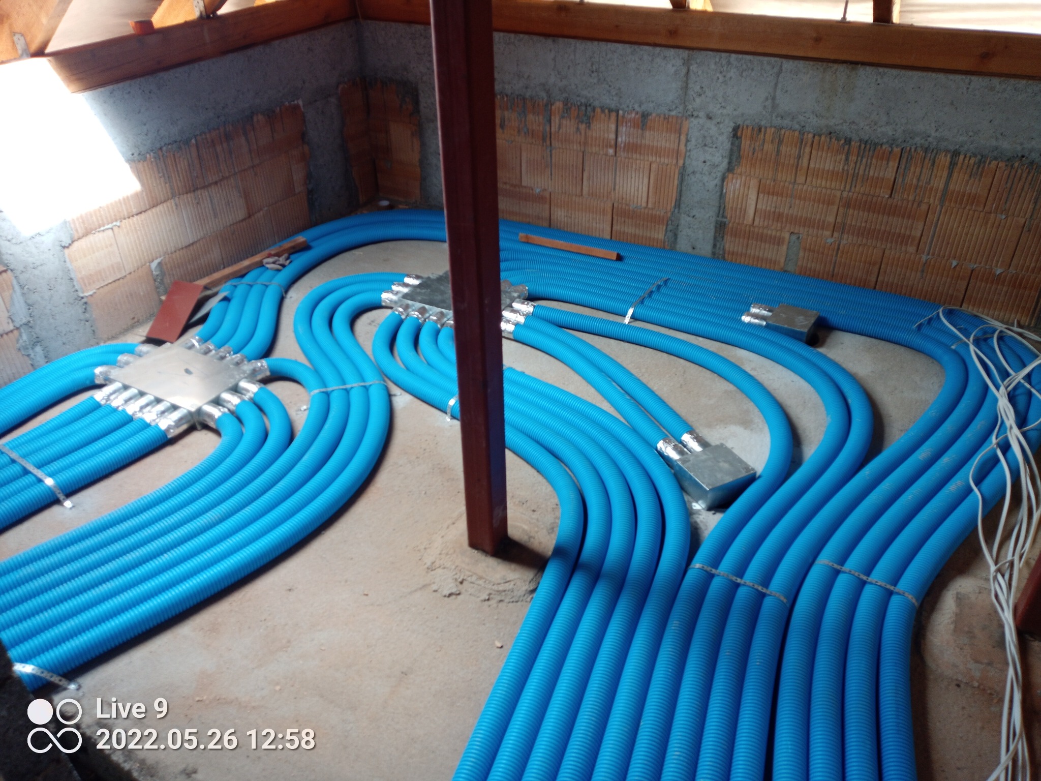 Niebieskie rury instalacji ogrzewania podłogowego, ułożone w pętle na surowym podłożu, z widocznymi elementami rozdzielczymi i fragmentem ściany z cegły.