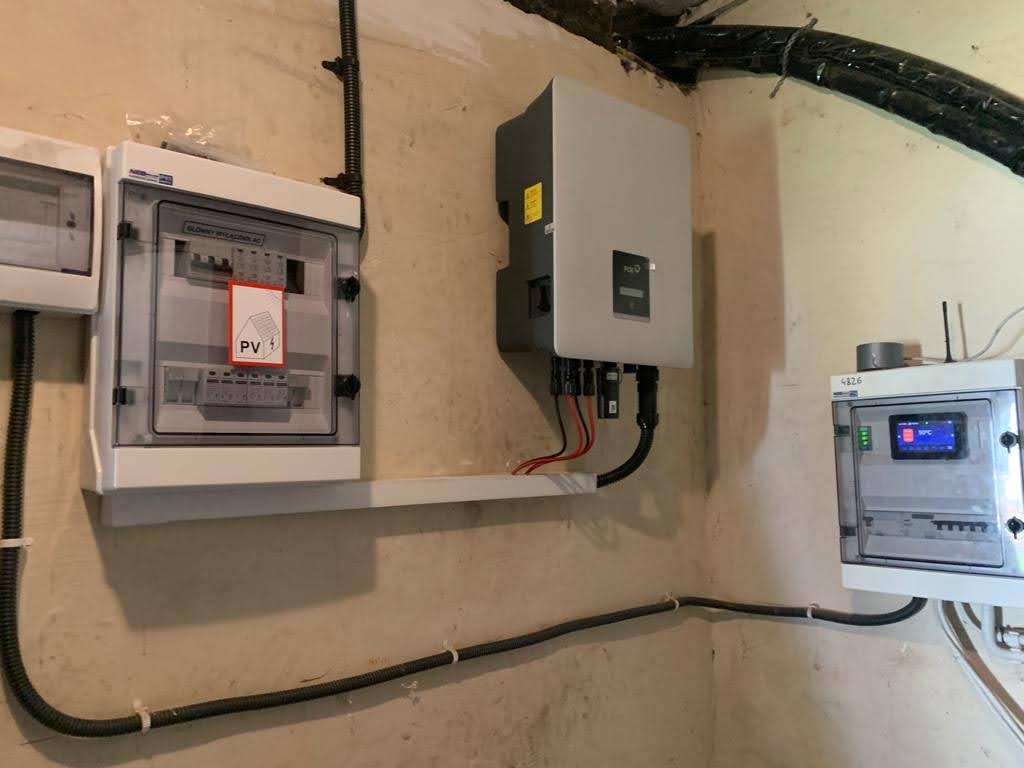 Instalacja elektryczna: rozdzielnice i inwerter fotowoltaiczny zamontowane na ścianie w pomieszczeniu technicznym. Widoczne przewody i elementy instalacji.