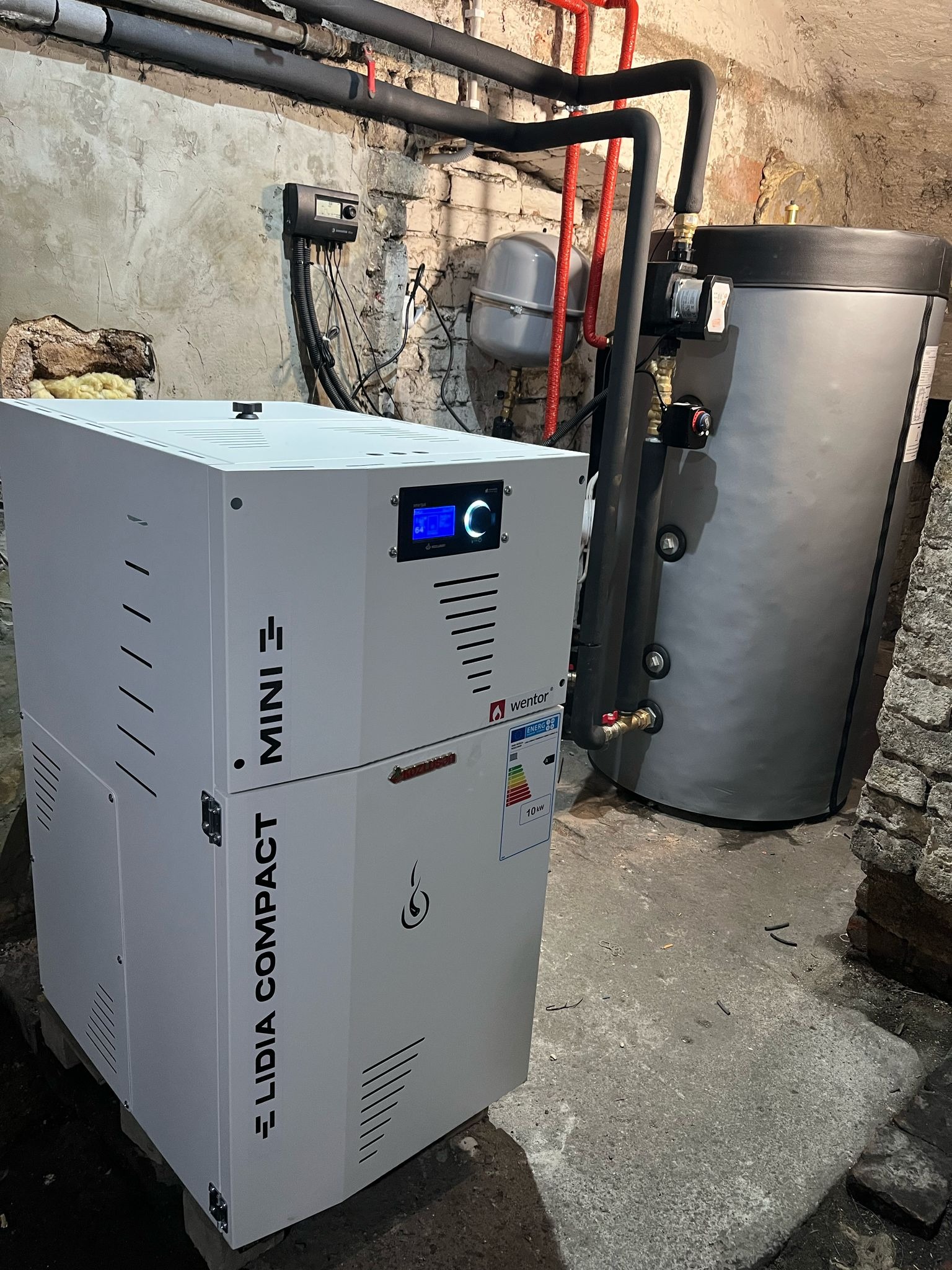 Kompaktowy piec gazowy Wentor Lidia Compact Mini z zasobnikiem w kotłowni o surowych ścianach. Widoczne rury instalacji CO i CWU.