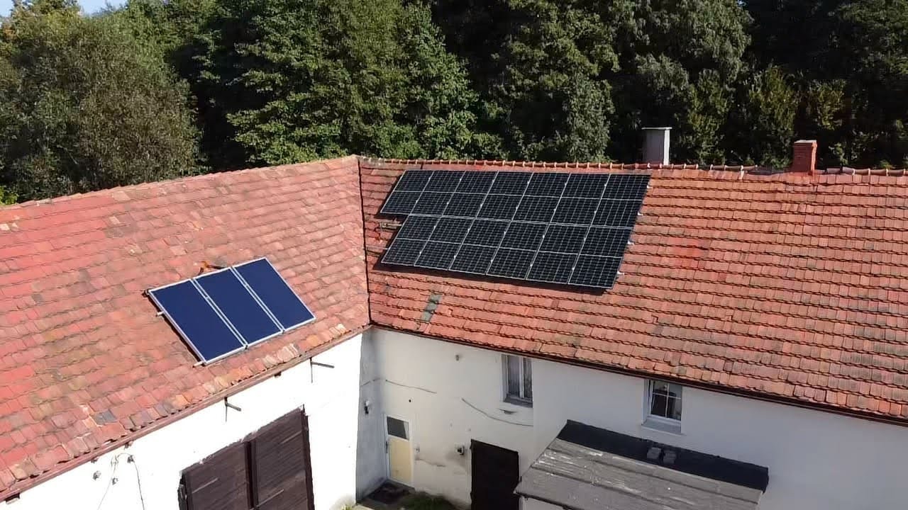 Instalacja paneli słonecznych na dachu budynku z czerwoną dachówką. Widok z góry, otoczenie zieleni. Dwa zestawy paneli: jeden większy, drugi mniejszy.