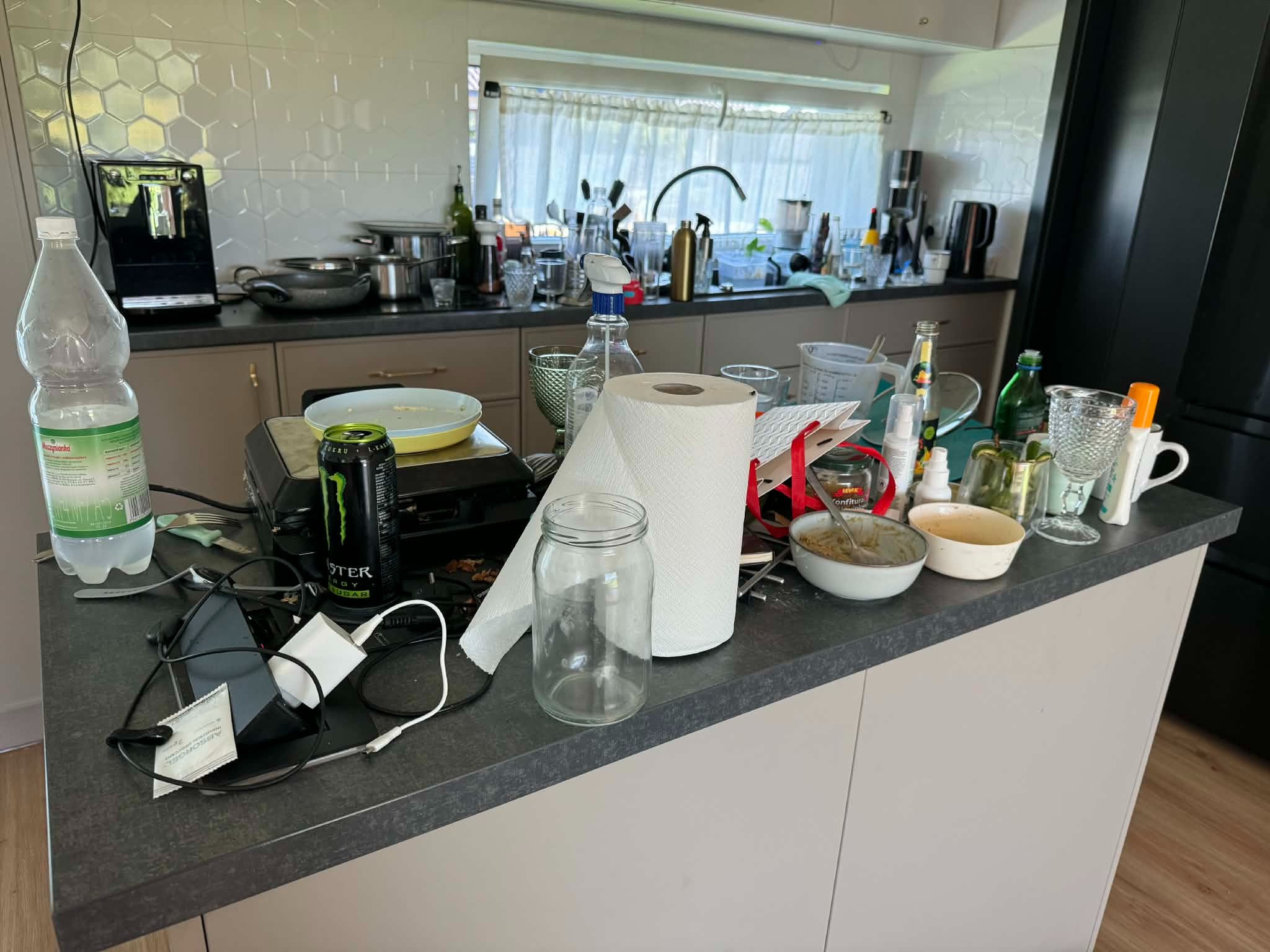 Zabałaganiony blat kuchenny z naczyniami, resztkami jedzenia, butelkami i akcesoriami kuchennymi. Widok na okno z firanką i zlewozmywak.