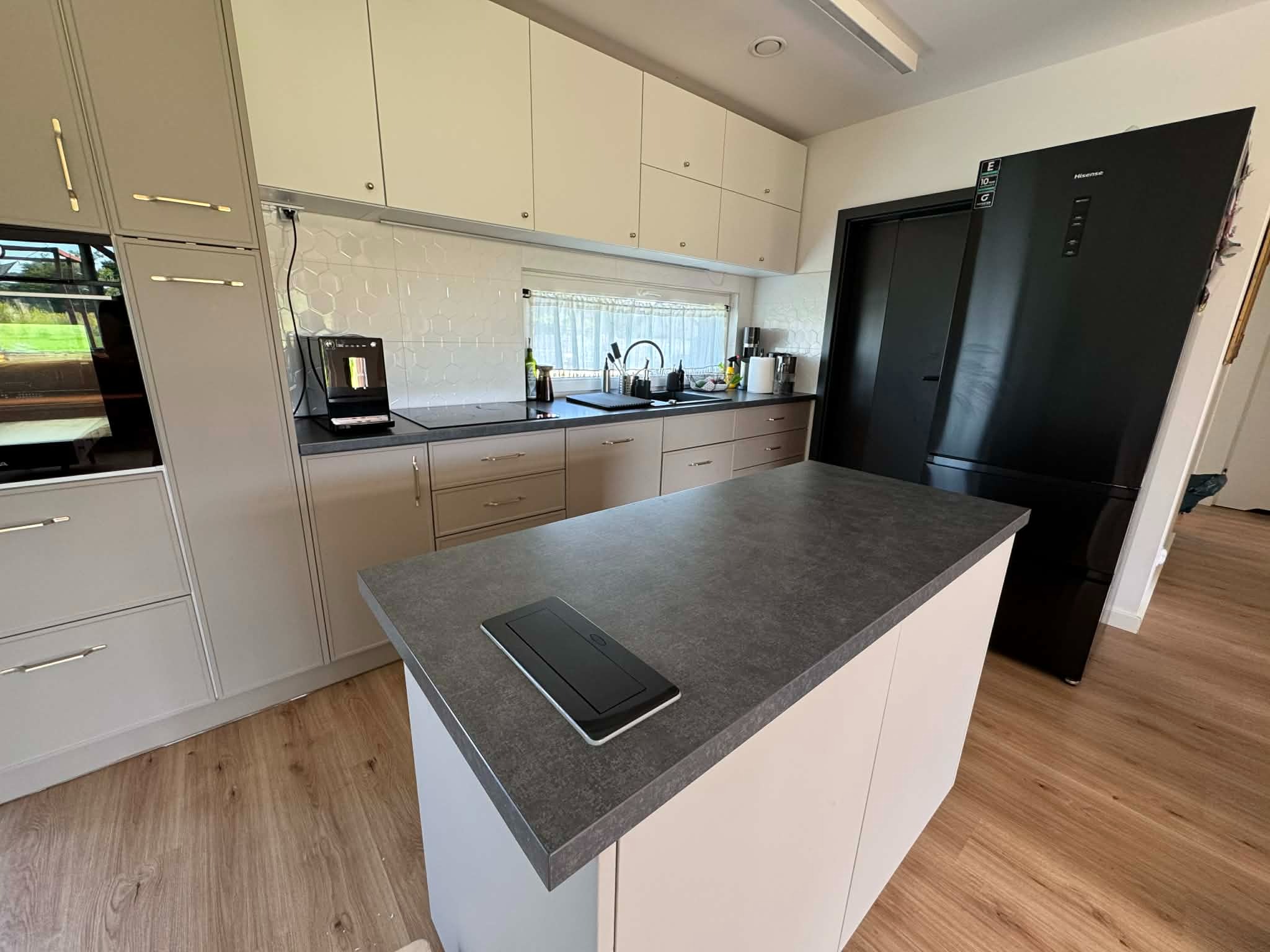 Jasna kuchnia z wyspą i czarną lodówką. Na blacie wyspy wbudowane gniazdko elektryczne. Białe płytki heksagonalne nad blatem. Drewniana podłoga.