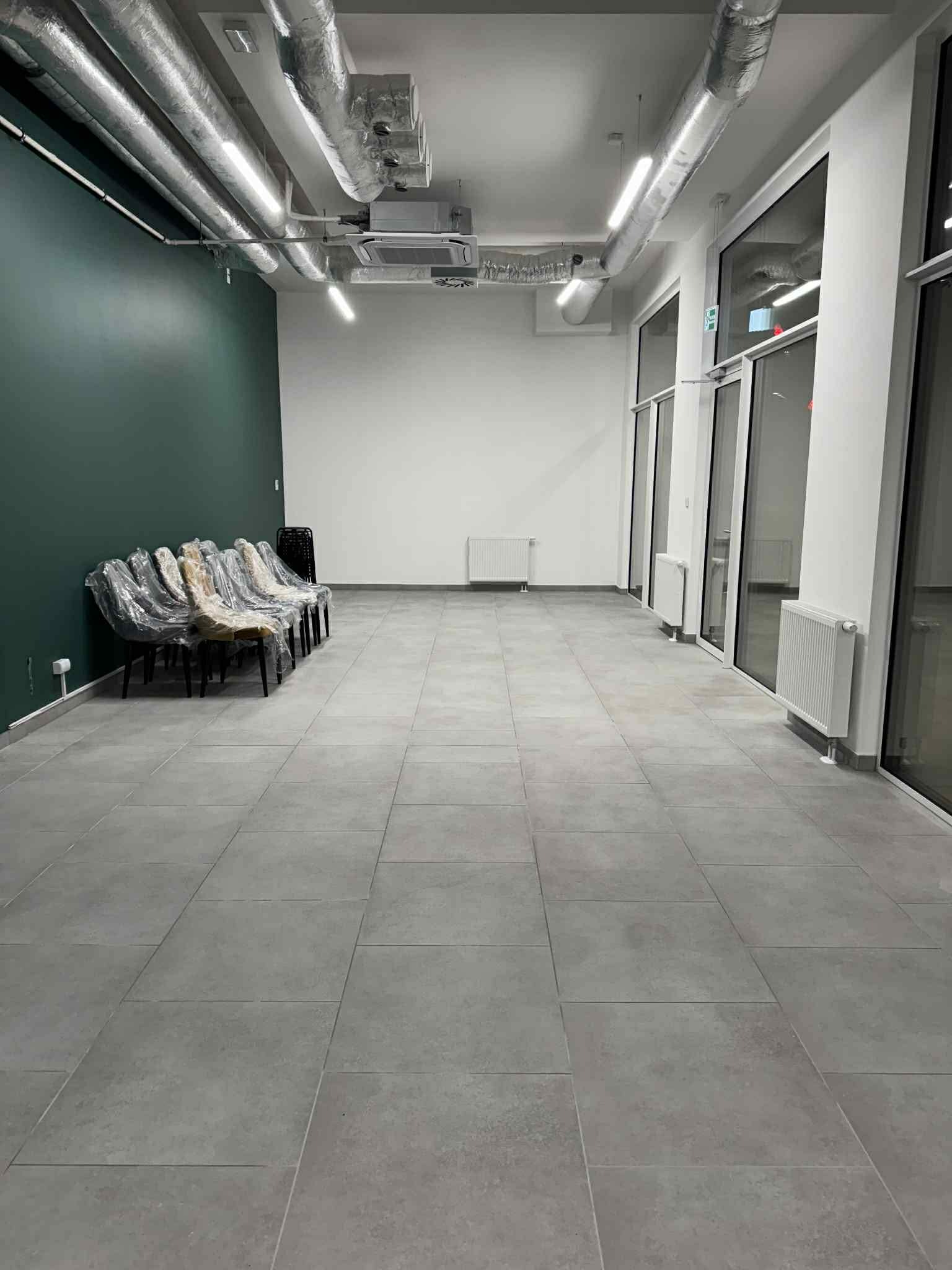 Wnętrze biurowe z szarymi płytkami podłogowymi, zieloną ścianą i rzędem krzeseł w folii ochronnej. Widoczne instalacje sufitowe i grzejniki.