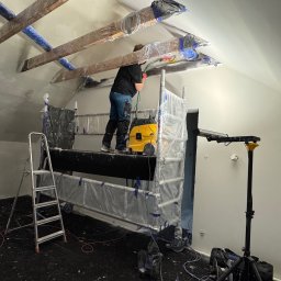 Budscar Budownictwo - Pracownik na rusztowaniu szlifuje sufit poddasza, zabezpieczone elementy drewniane, odkurzacz przemysłowy, przygotowanie do malowania, widok z dołu.