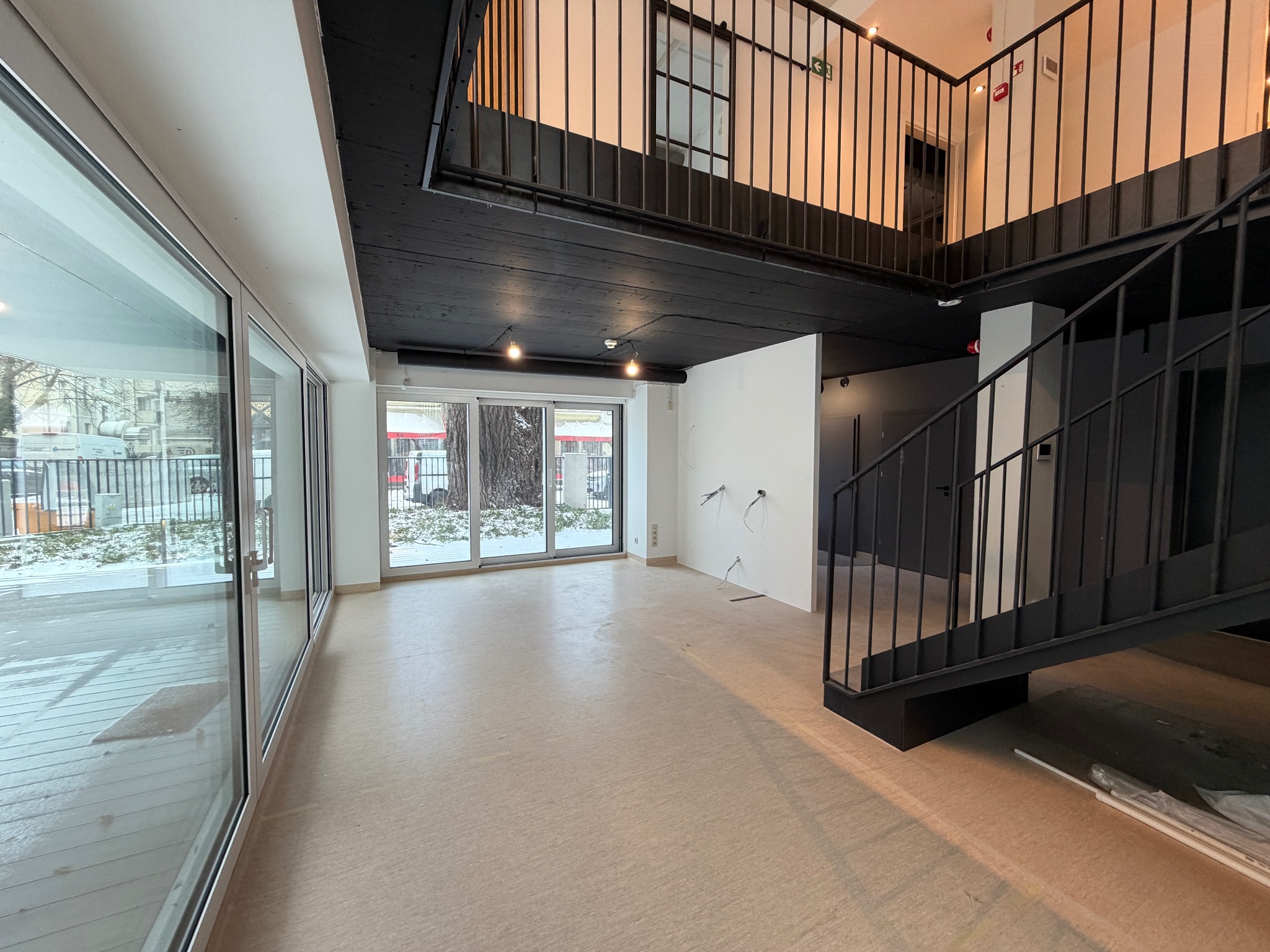 Przestronne, jasne wnętrze w stylu loft z dużymi oknami i antresolą. Widoczne surowe wykończenie, instalacje elektryczne i schody z metalową balustradą. Podłoga wyłożona panelami.