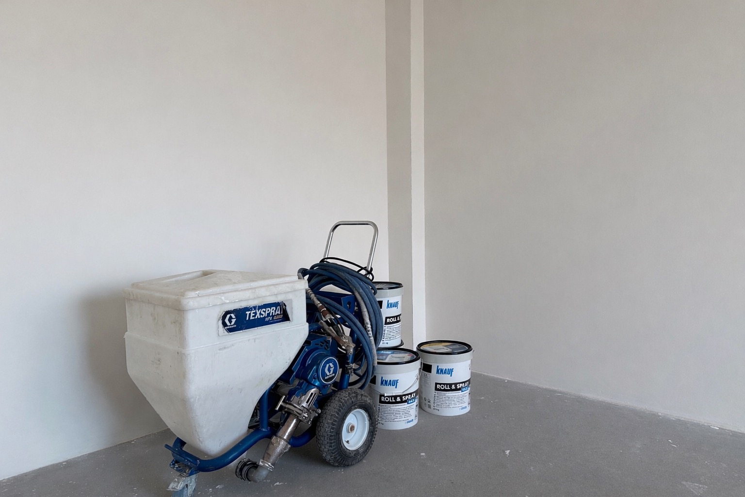 Biała ściana po malowaniu natryskowym, widoczny agregat malarski TexSpray i puszki z farbą Knauf Roll & Spray. Minimalistyczne ujęcie, neutralne światło.