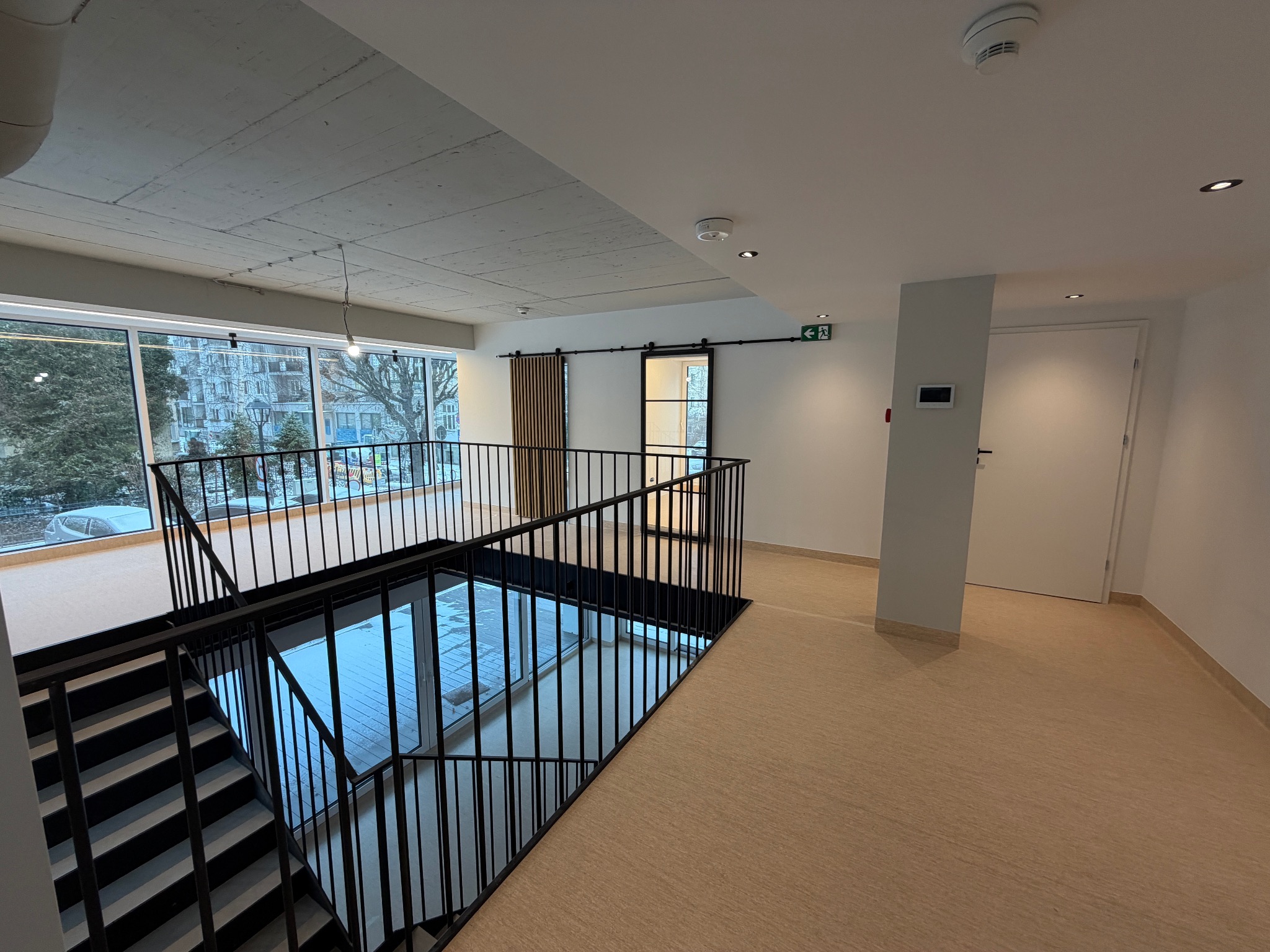 Nowoczesne wnętrze z betonowym sufitem, dużymi oknami i czarną balustradą schodów. Widoczne drzwi przesuwne i jasna podłoga. Minimalistyczny design.