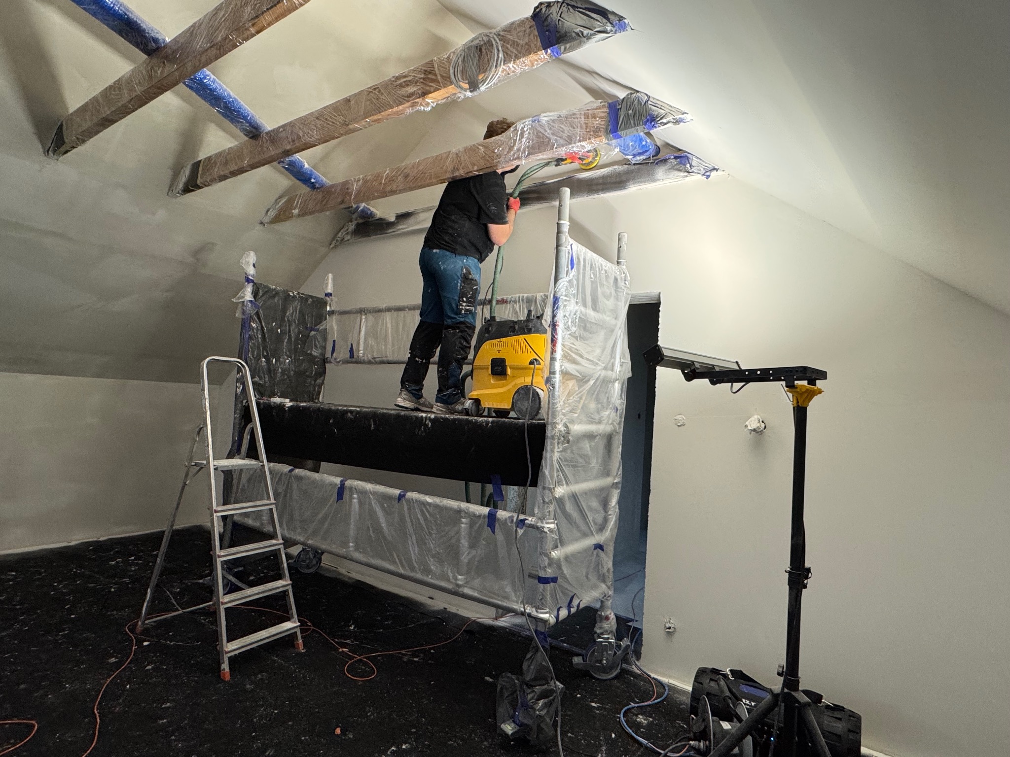 Pracownik na rusztowaniu szlifuje sufit poddasza, zabezpieczone elementy drewniane, odkurzacz przemysłowy, przygotowanie do malowania, widok z dołu.