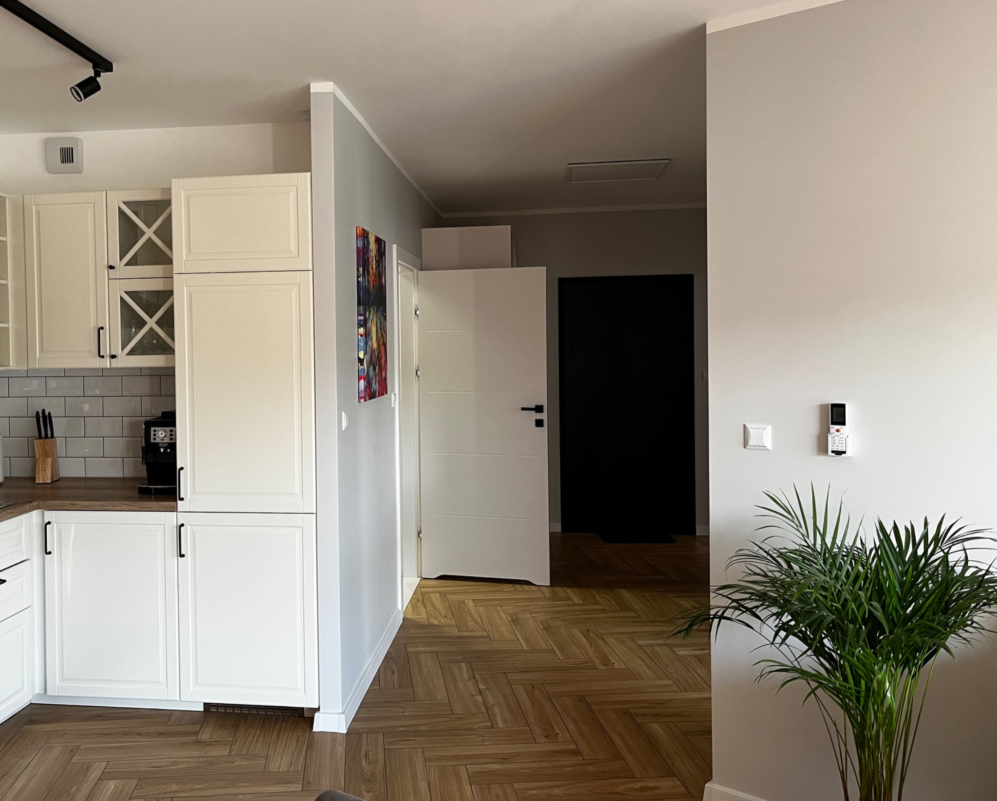 Jasne wnętrze: biała zabudowa kuchenna z drewnianym blatem, parkiet w jodełkę, szara ściana z rośliną doniczkową i włącznikiem. Nowoczesny design.