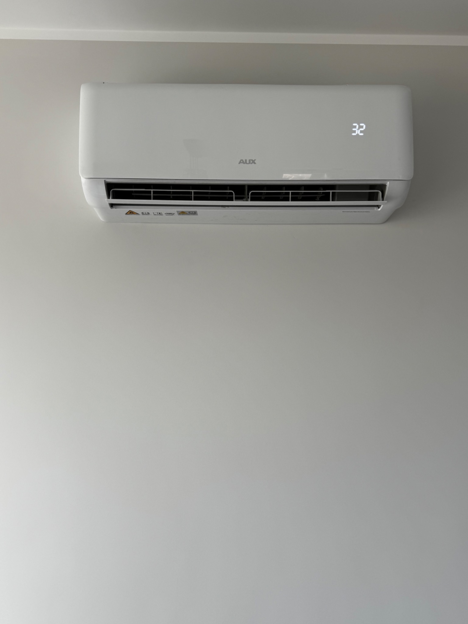 Biały klimatyzator AUX zamontowany na jasnej ścianie, wyświetlający temperaturę 32 stopnie. Widoczne detale urządzenia i otoczenia.