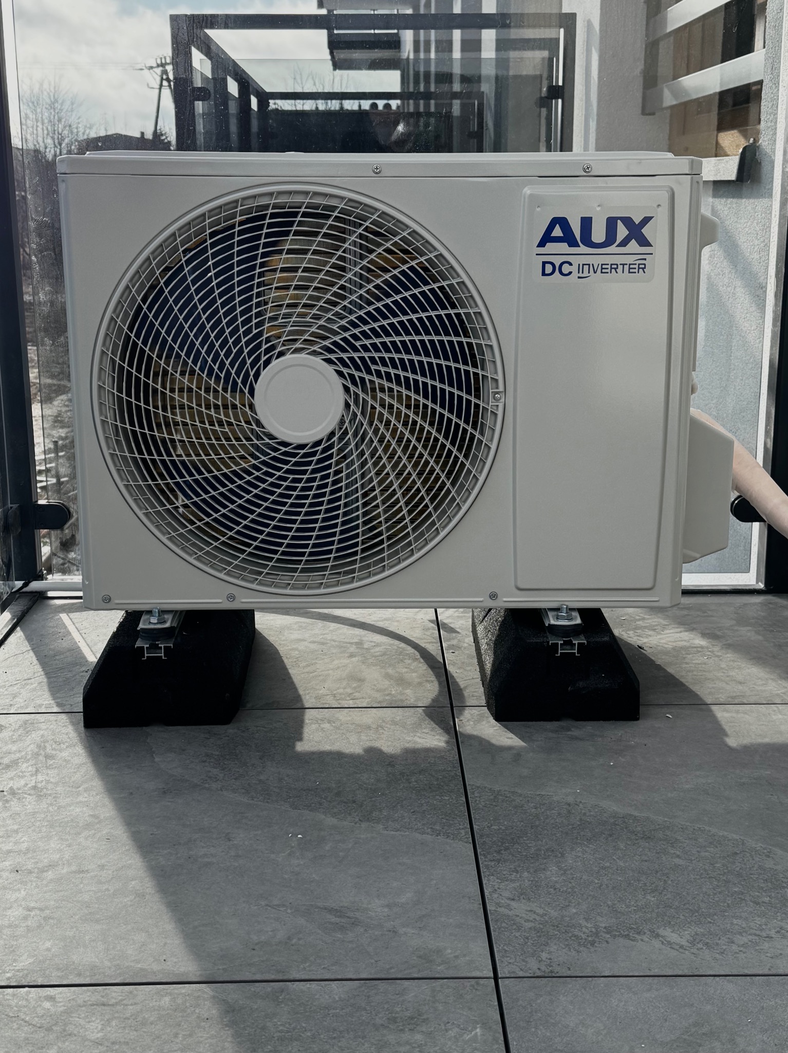 Biała jednostka zewnętrzna klimatyzacji AUX DC Inverter, zamontowana na czarnych podstawach na szarym tarasie, widok z dołu, z elementami otoczenia w tle.