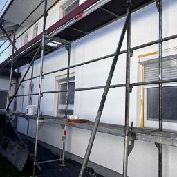 Tomasz Skwirzynski - Biała elewacja domu w trakcie odnawiania, widoczna z poziomu rusztowania. Okna zabezpieczone folią, w tle góry.