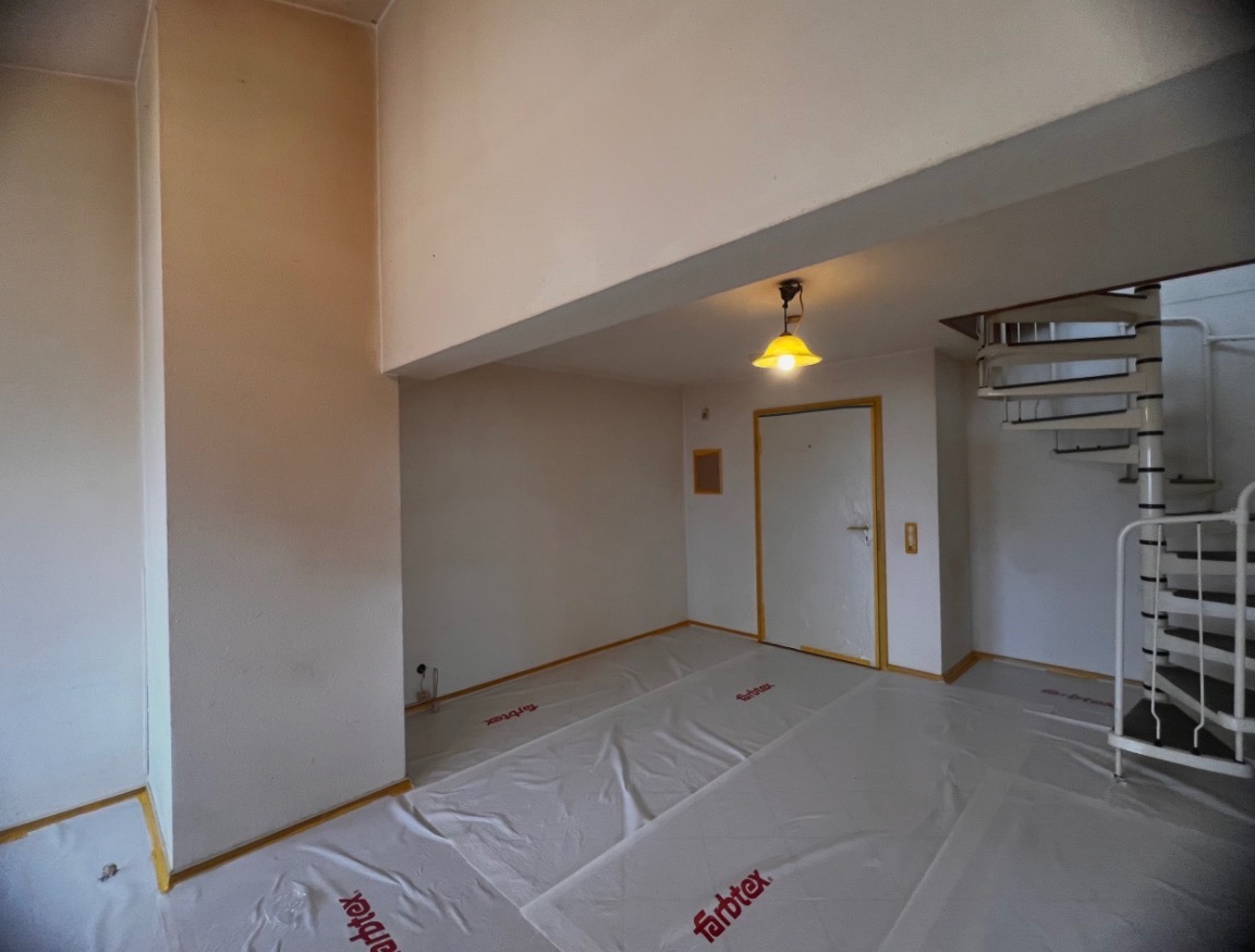 Przygotowane do malowania wnętrze z spiralnymi schodami. Podłoga zabezpieczona folią malarską, ściany w jasnym odcieniu, widoczna lampa i drzwi z żółtą ramą.