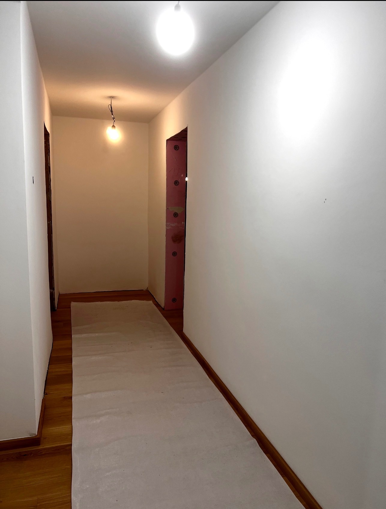 Korytarz po świeżym malowaniu na biało, zabezpieczona podłoga, widoczne ślady po remoncie. Oświetlenie sufitowe. Ujęcie z dołu, perspektywa wnętrza.