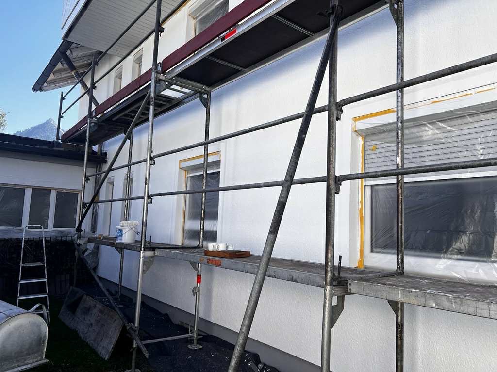 Biała elewacja domu w trakcie odnawiania, widoczna z poziomu rusztowania. Okna zabezpieczone folią, w tle góry.