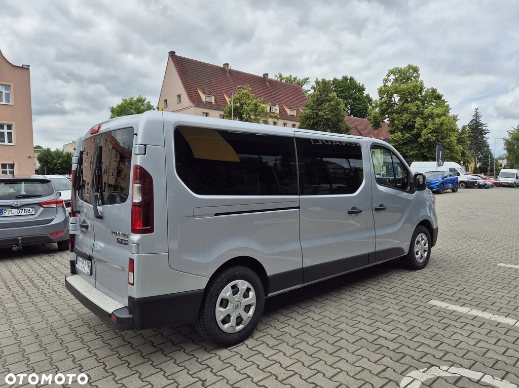 Srebrny bus Renault Trafic z przyciemnianymi szybami, zaparkowany na brukowanym placu, widok z tyłu i boku, w tle budynki i inne pojazdy.