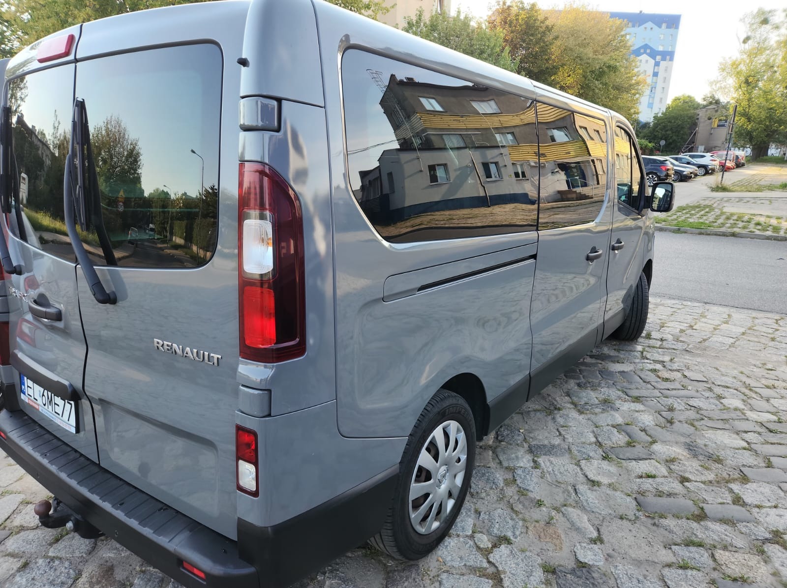 Szary Renault Trafic z widocznym numerem rejestracyjnym, zaparkowany na brukowanej ulicy. W oknach odbija się budynek.