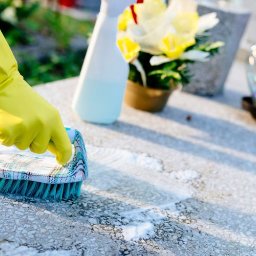 Promar kompleksowe usługi sprzątające - Czyszczenie nagrobka szczotką i detergentem w żółtych rękawicach ochronnych. W tle kwiaty i lampion.