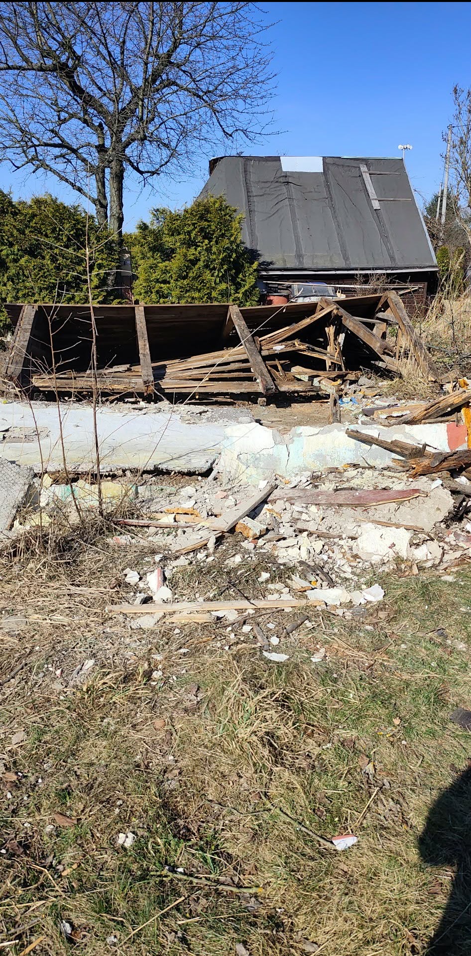 Zdemolowany budynek z gruzem i drewnianymi elementami na pierwszym planie, zarośnięty teren, drzewa w tle i fragment nieba. Widok na pozostałości po rozbiórce.