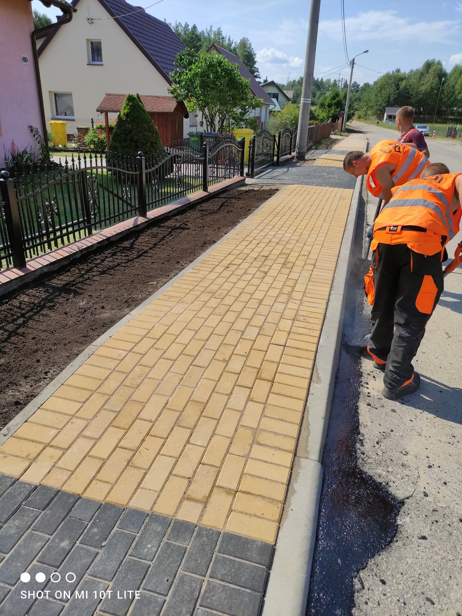 Żółta kostka brukowa ułożona wzdłuż chodnika, obok świeżo nasypana ziemia pod ogrodzeniem. Pracownicy w pomarańczowych kamizelkach układają obrzeża.