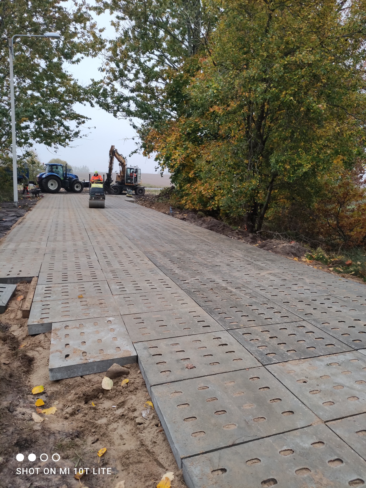 Układanie ażurowych płyt betonowych na gruncie. W tle maszyny budowlane: traktor, walec i koparka. Prace ziemne w toku, widoczne jesienne liście.