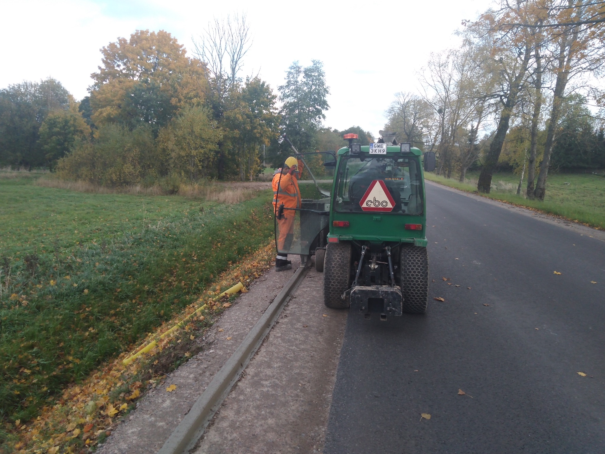 Pracownik w stroju roboczym przy zielonym pojeździe specjalnym na poboczu drogi, prawdopodobnie podczas prac ziemnych lub instalacyjnych. Widoczne żółte rury.