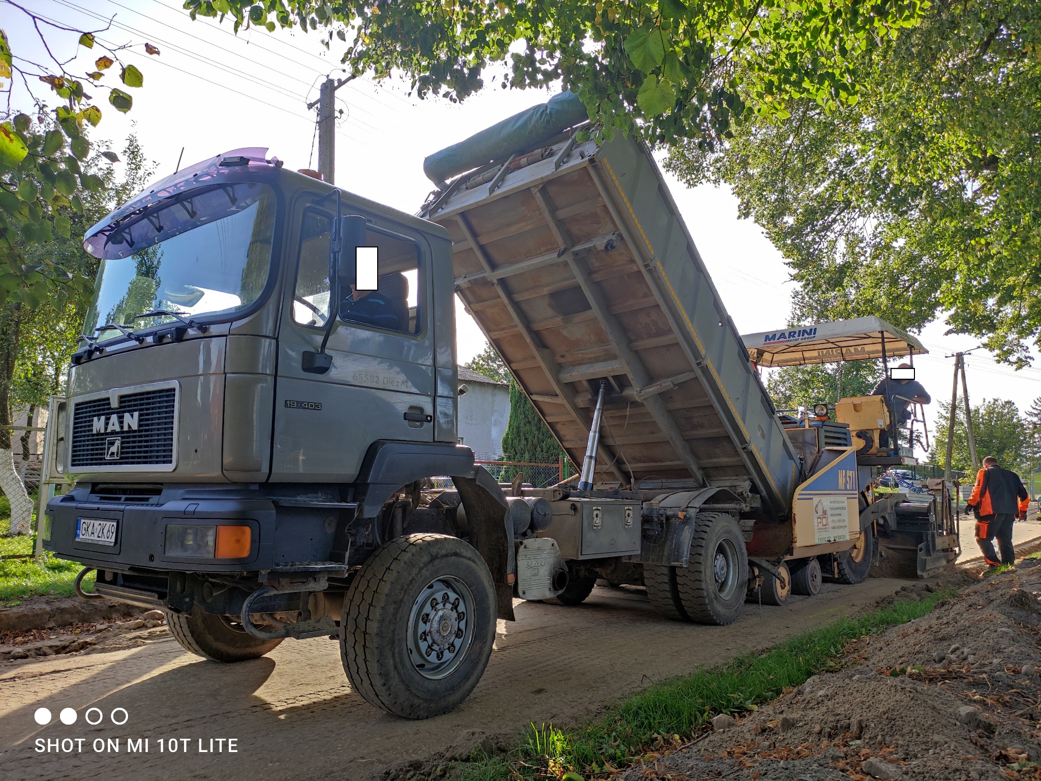 Ciężarówka MAN wyładowuje materiał do rozściełacza asfaltu Marini na drodze w słoneczny dzień. Widoczny pracownik w odblaskowej odzieży. Prace drogowe w toku.