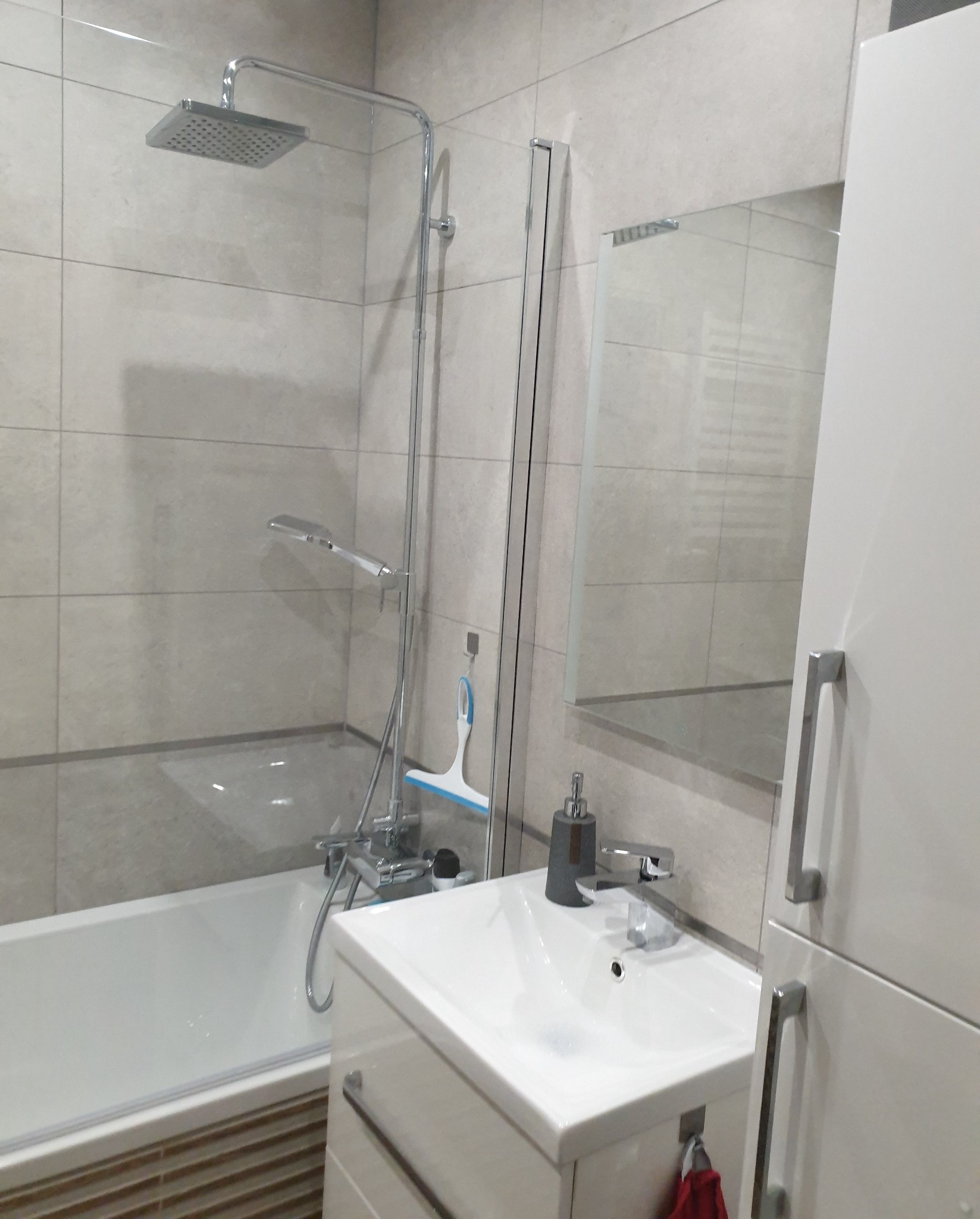 Jasna łazienka z wanną i kabiną prysznicową typu walk-in, umywalką z szafką i lustrem. Na ścianach płytki w odcieniach beżu. Widoczny dozownik do mydła i ściągaczka do szyb.