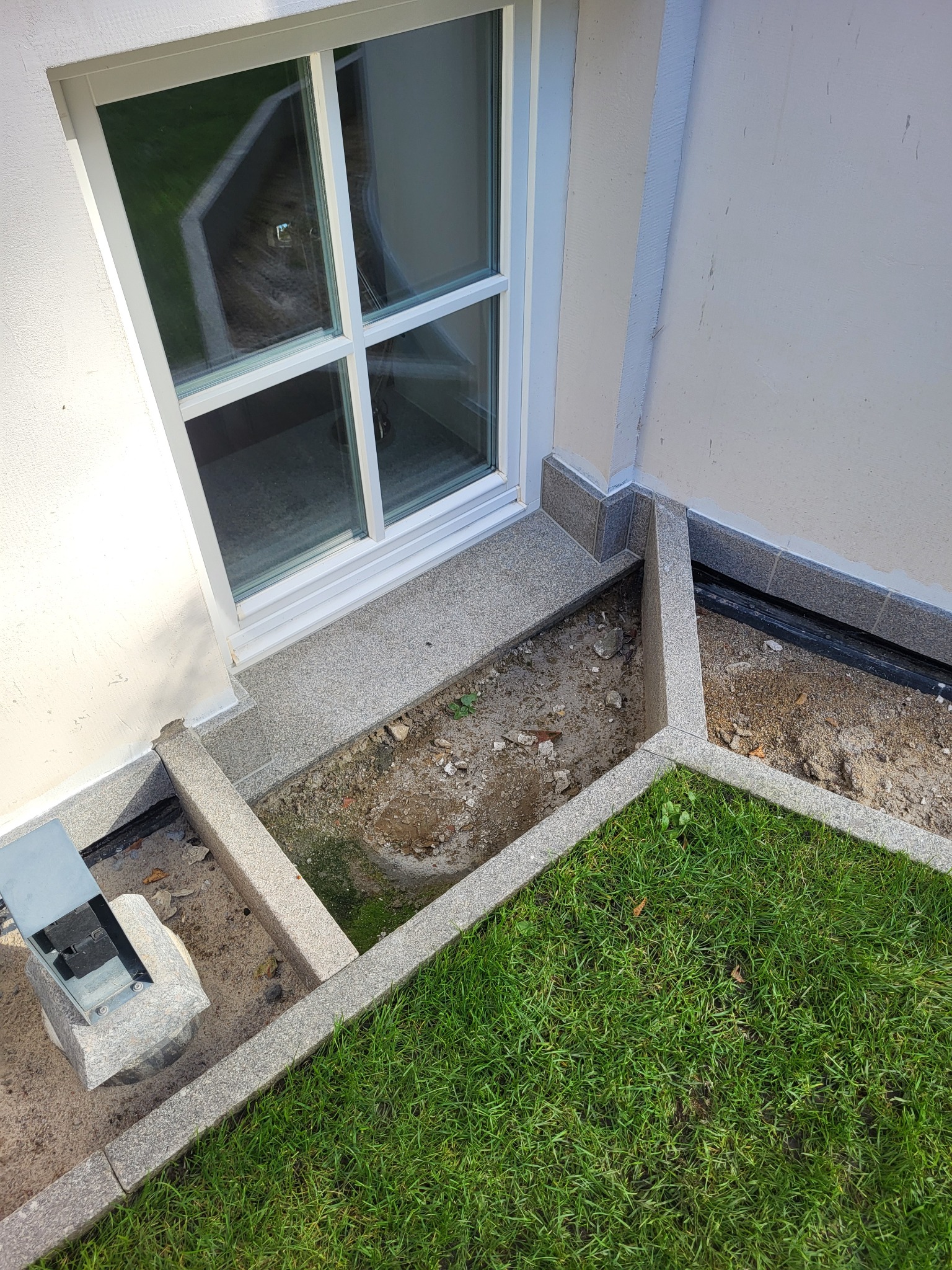 Wykończenie okna piwnicznego z szarego granitu, z widoczną instalacją odwadniającą i fragmentem trawnika. Detal architektoniczny.