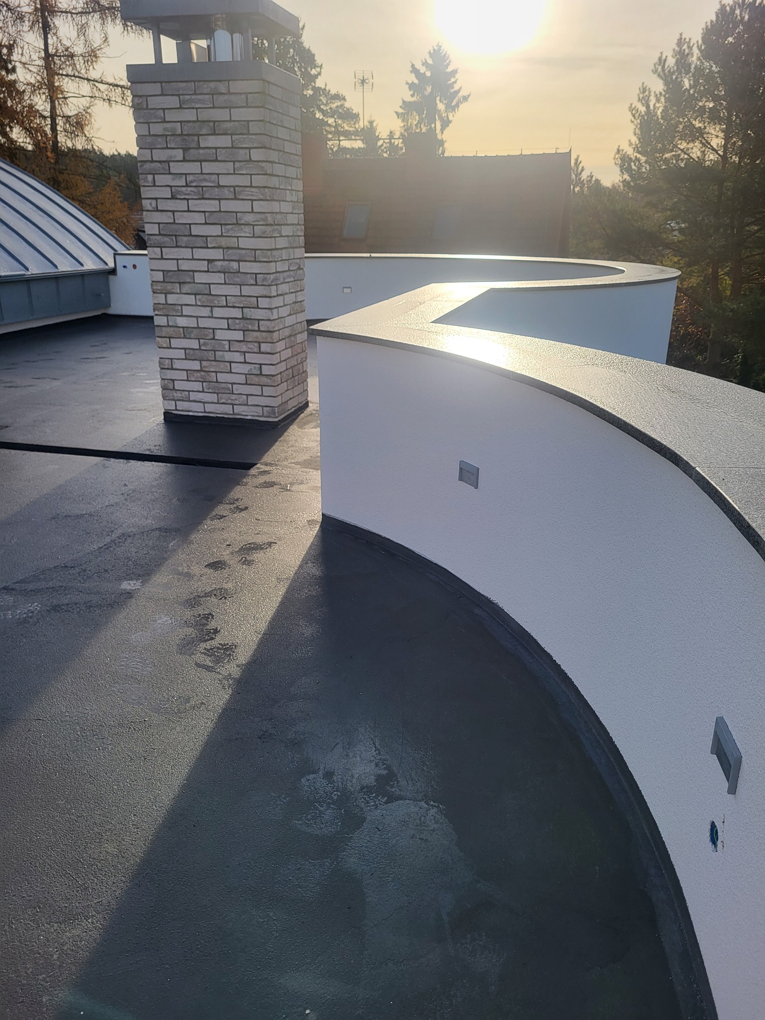 Wykończony dach płaski z ceglanym kominem i białym, zaokrąglonym murem. Ślady stóp na powierzchni dachu. Słoneczne oświetlenie, widok z góry.