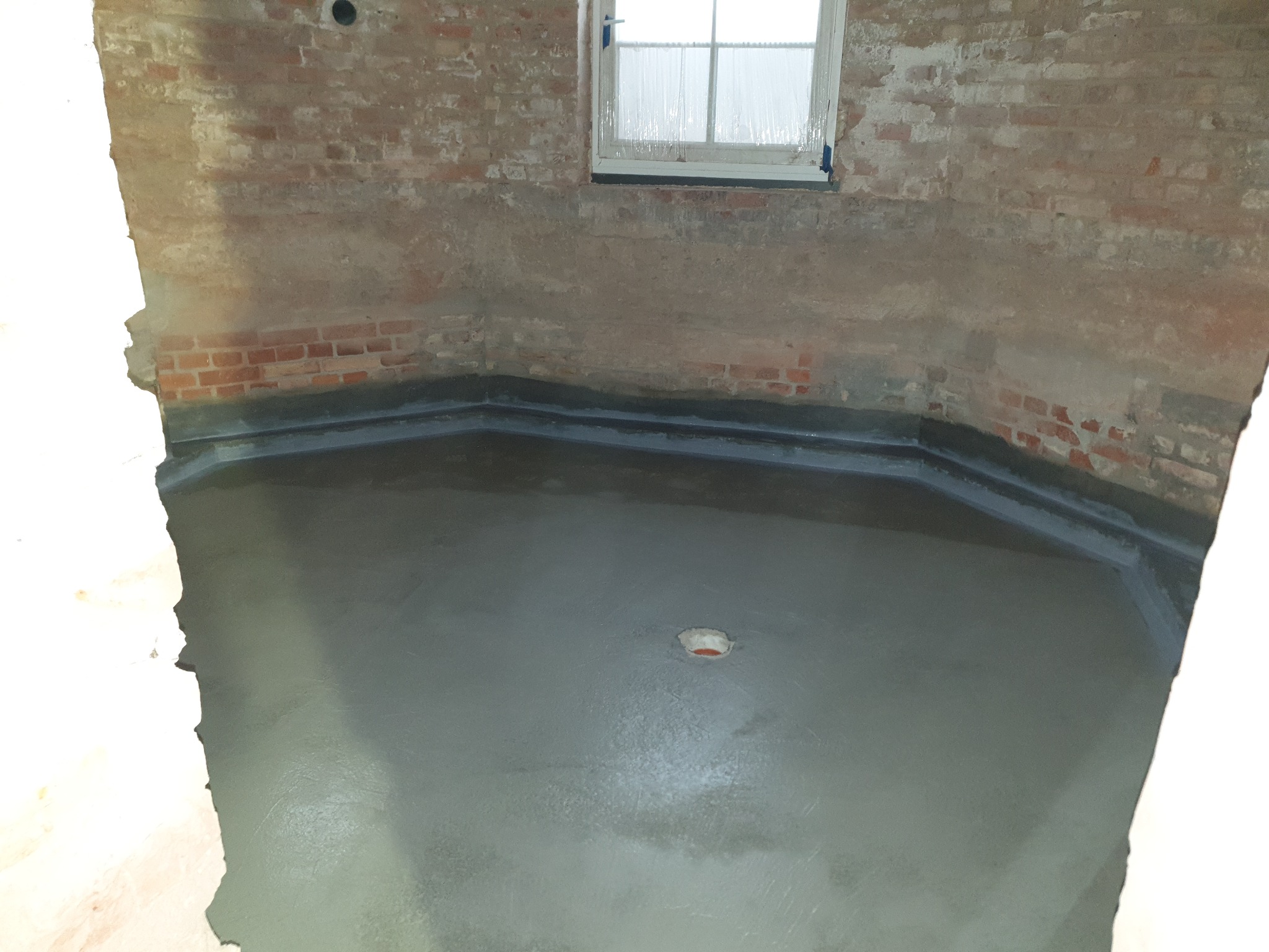 Wylewka podłogowa w łazience z surowymi, ceglanymi ścianami i oknem. Widoczny otwór odpływowy. Etap prac wykończeniowych w pomieszczeniu o nieregularnym kształcie.