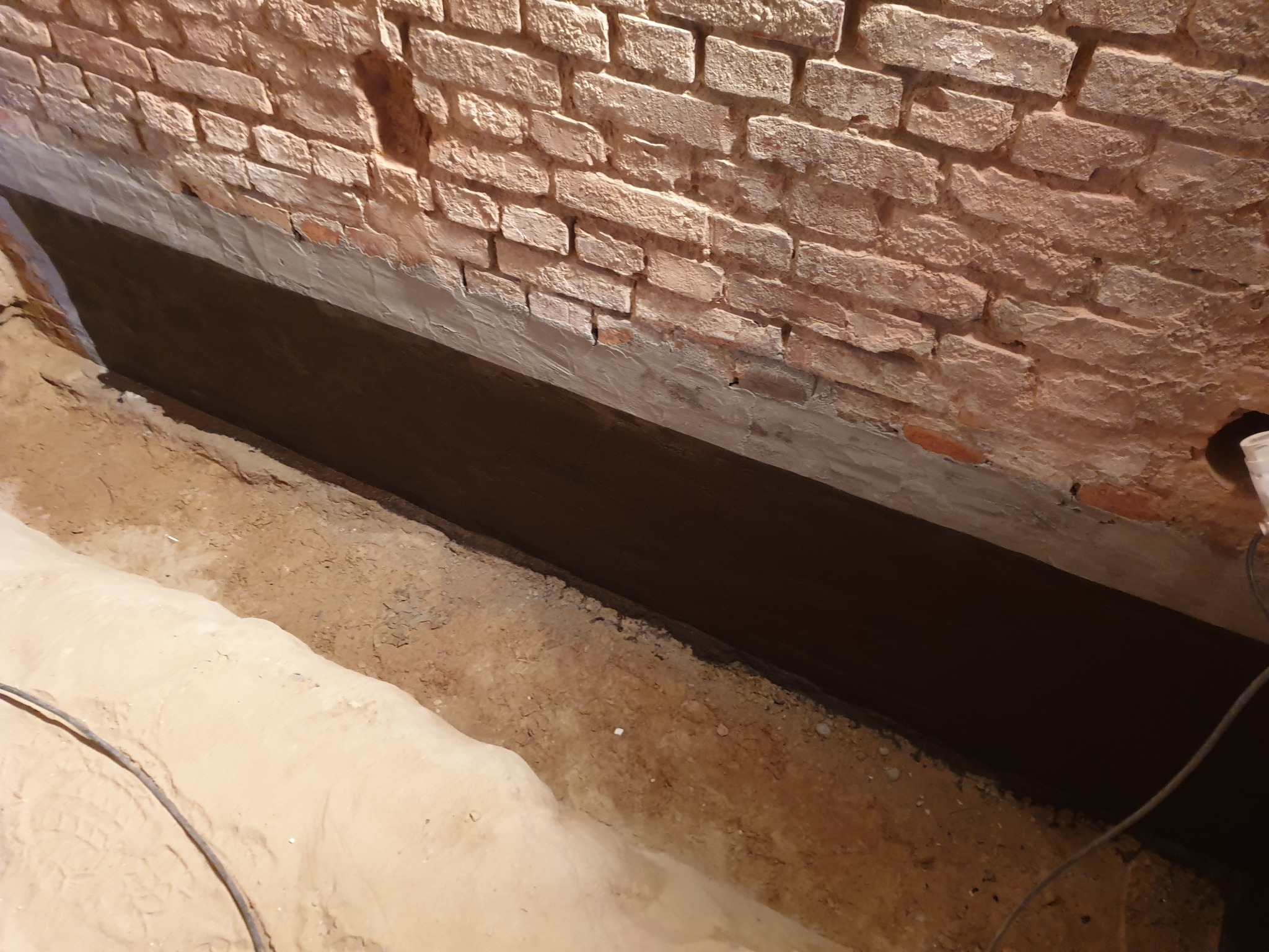 Hydroizolacja fundamentów z cegły, zabezpieczenie przed wilgocią. Widoczne warstwy izolacyjne i grunt. Przewód elektryczny w rogu kadru.