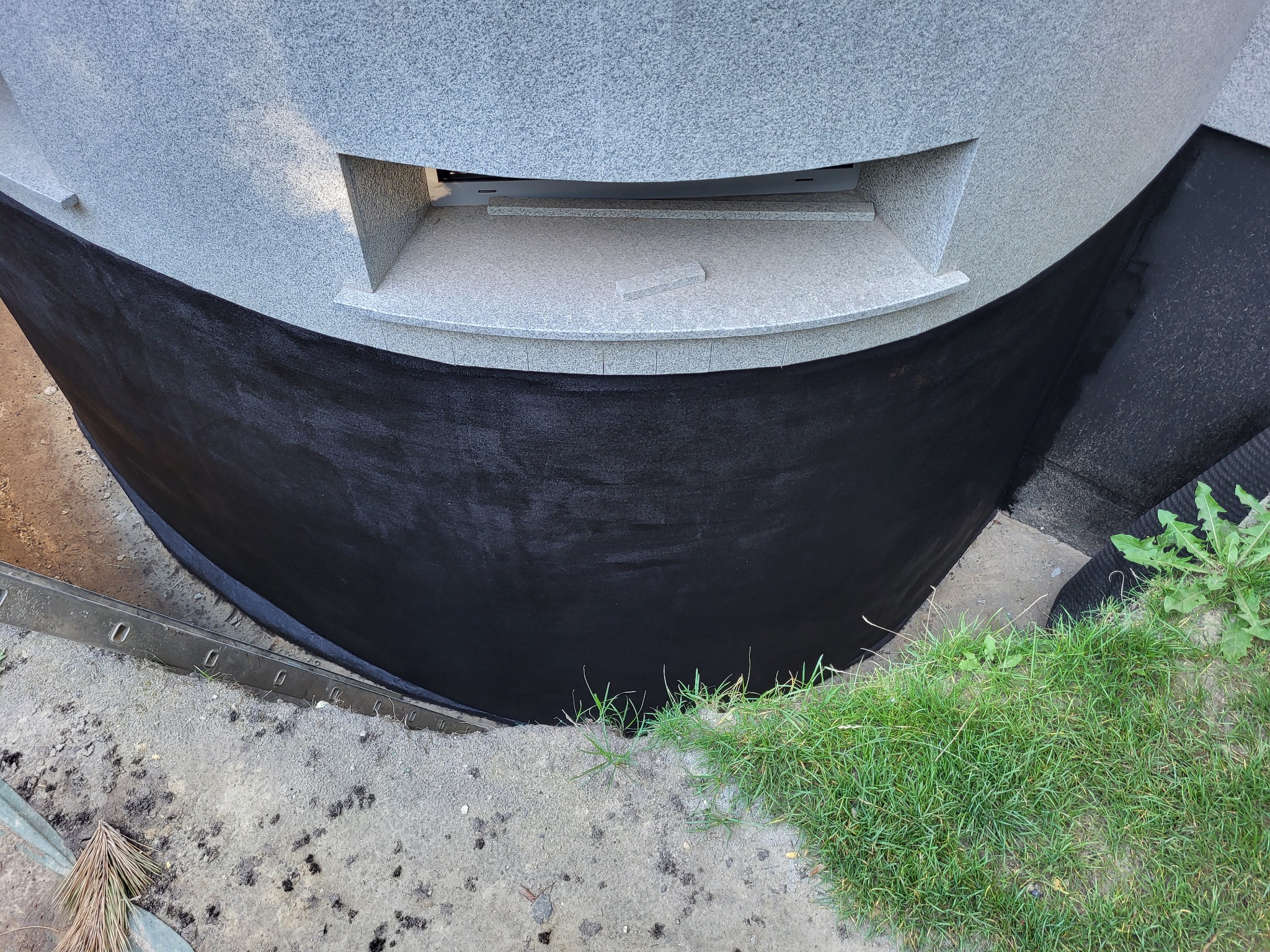 Hydroizolacja fundamentów z szarego granitu, zabezpieczona czarną powłoką bitumiczną. Widoczne detale wykończenia i fragment zieleni przy podstawie.