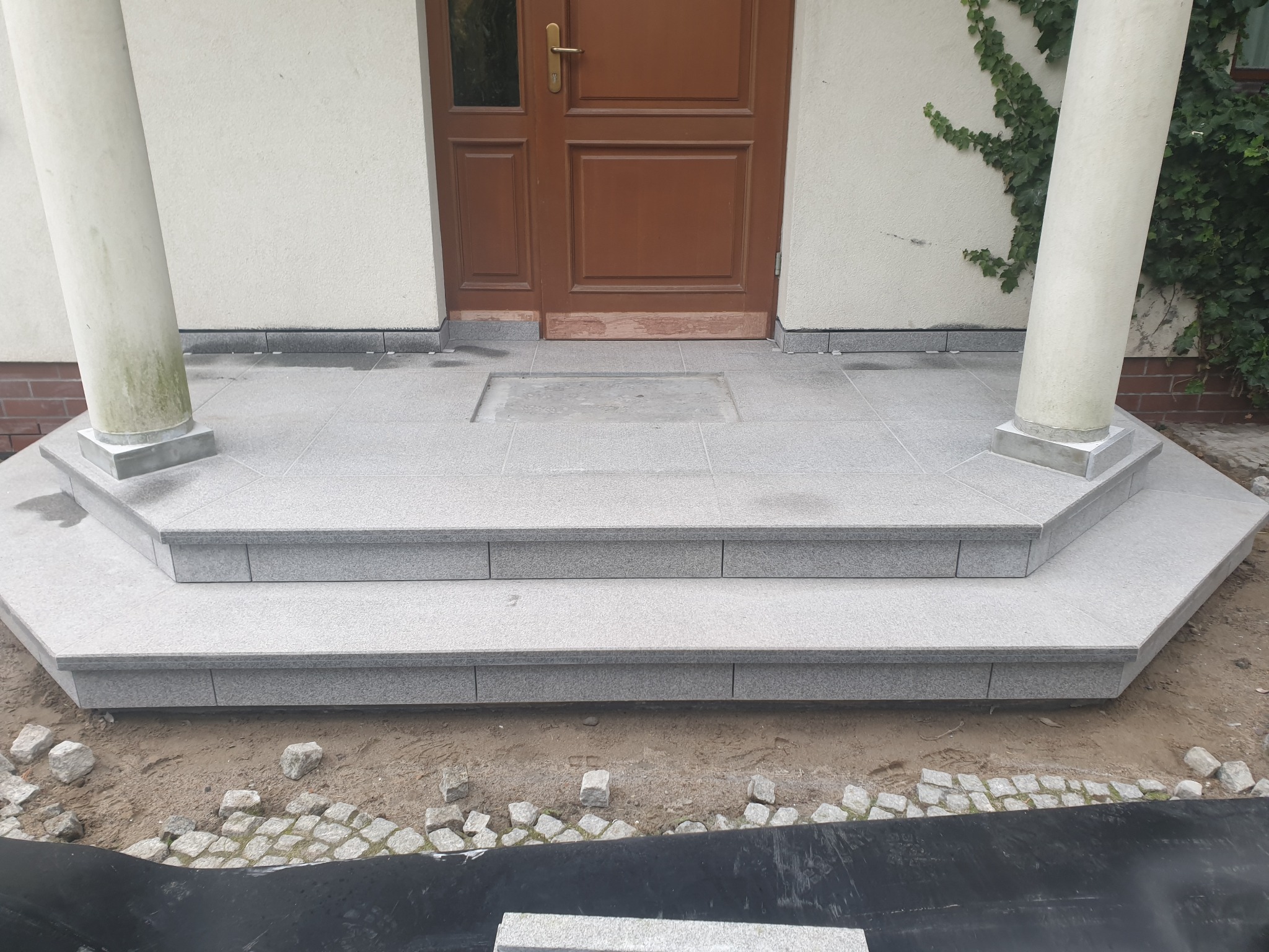 Granitowe schody wejściowe z kolumnami, brązowe drzwi, jasna elewacja z bluszczem. Wykończenie zewnętrzne domu, widok z dołu, kostka brukowa w tle.