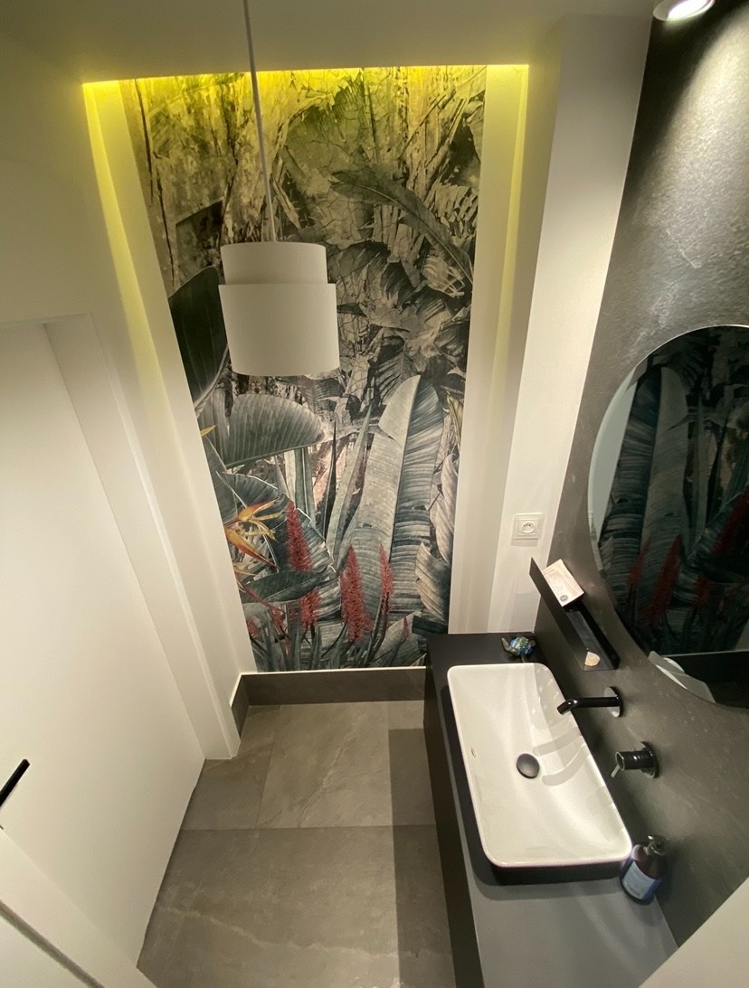 Nowoczesna łazienka z umywalką nablatową, okrągłym lustrem i tapetą w tropikalne liście, oświetlona lampą wiszącą i listwą LED.