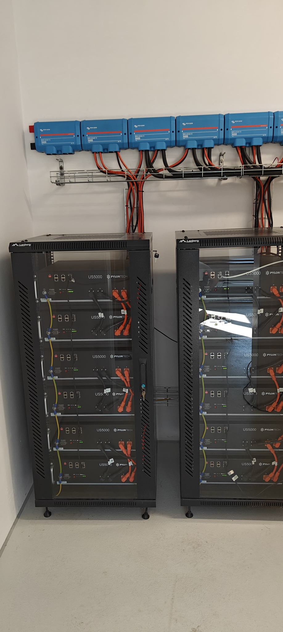 Dwa regały Lanberg z bateriami Pylontech US5000, połączone przewodami z niebieskimi inwerterami na ścianie. Instalacja magazynu energii w pomieszczeniu.