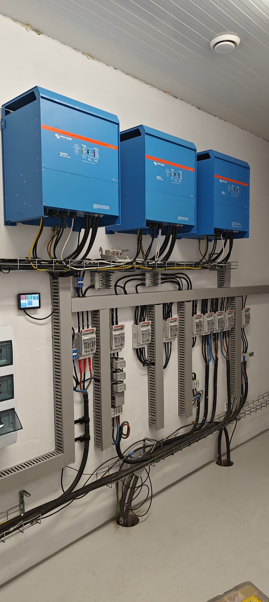 Instalacja trzech niebieskich inwerterów Victron Energy na ścianie, z widocznymi kablami i zabezpieczeniami w metalowej korycie. Panel sterowania po lewej stronie.